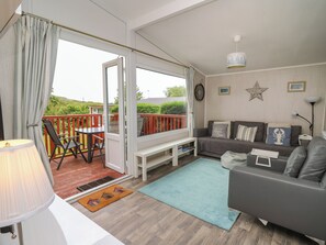 A cozy living room with a sofa, a coffee table, and a dining area with a table and chairs. There is a balcony with a railing and a view of greenery outside. - Chalet 64 (Tywyn)