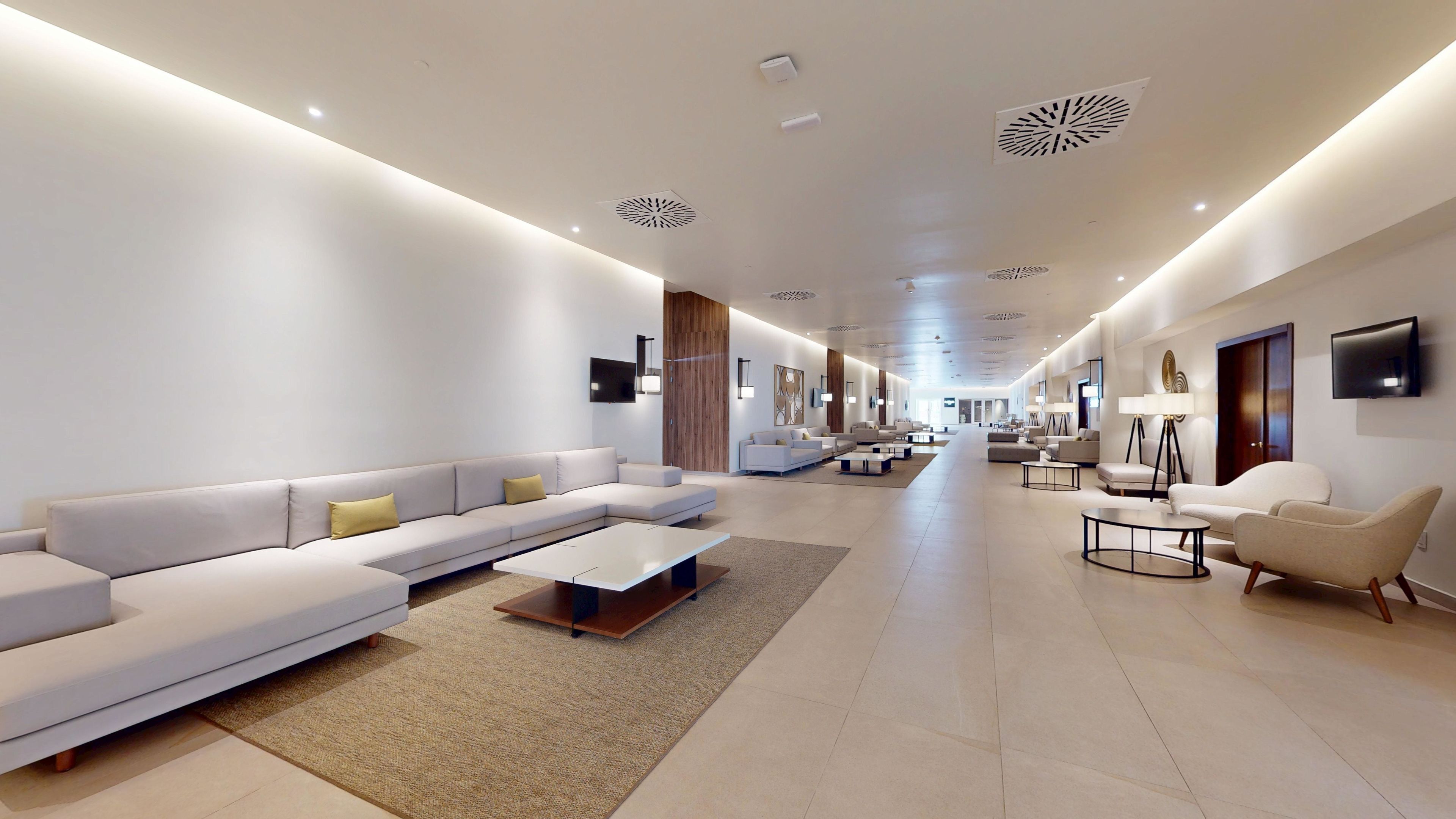 A spacious, modern hotel lobby with a long reception area, multiple seating options including sofas and armchairs, and a minimalist design with neutral colors.