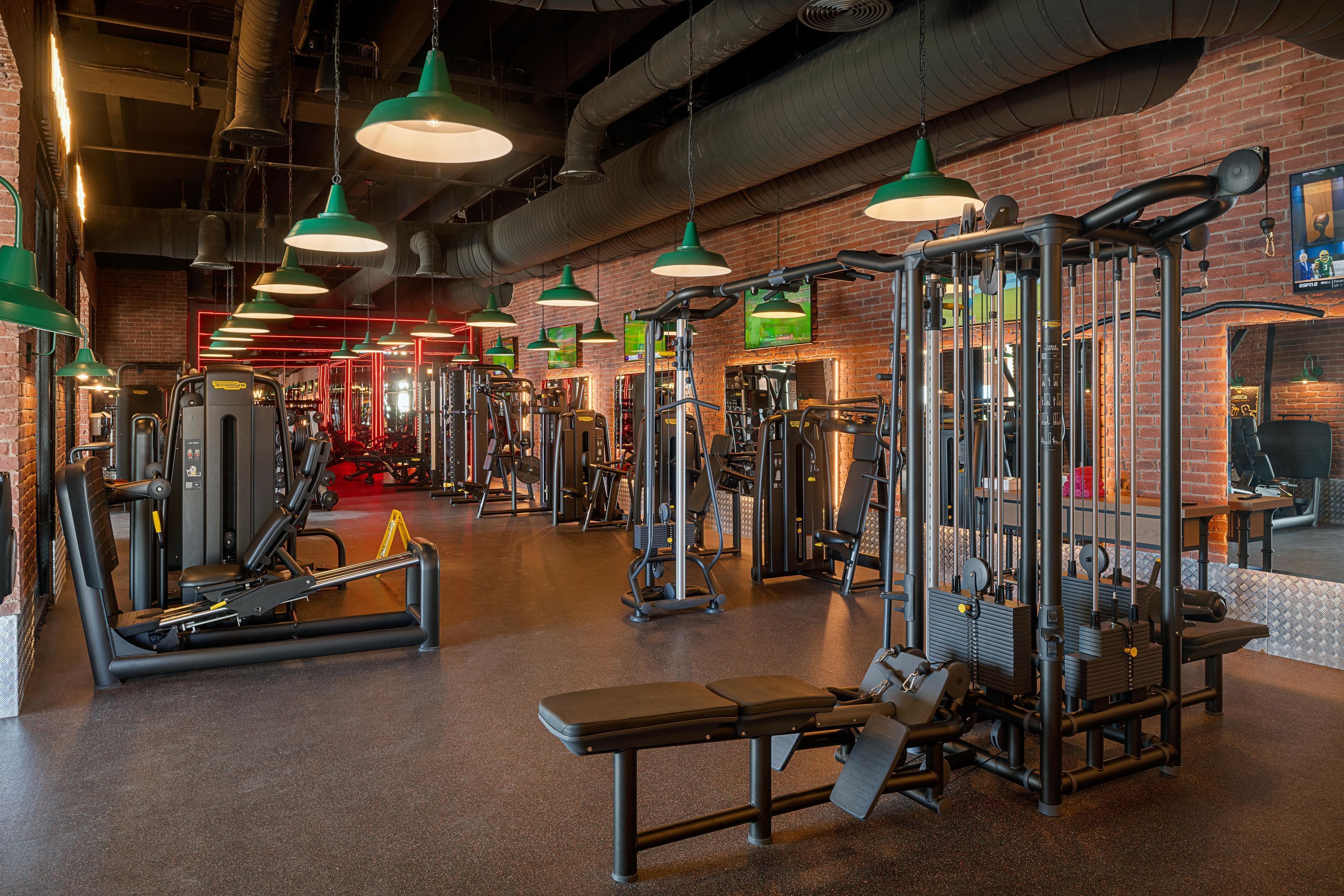 Een sportschool met diverse fitnessapparaten, groene hanglampen en een bakstenen muur.