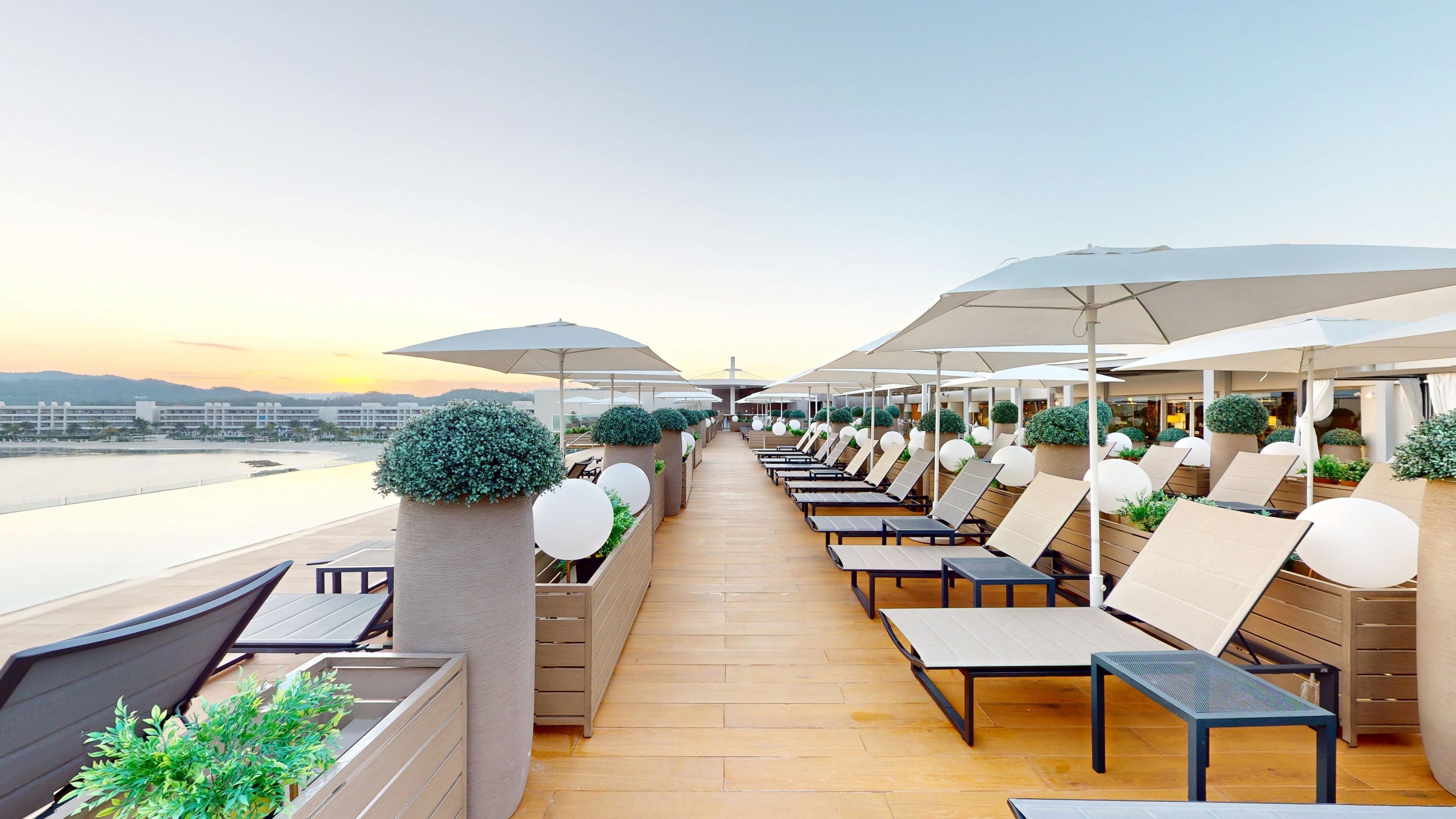 Terraza con sillones de descanso y sombrillas, con vista a un cuerpo de agua y edificios al fondo.