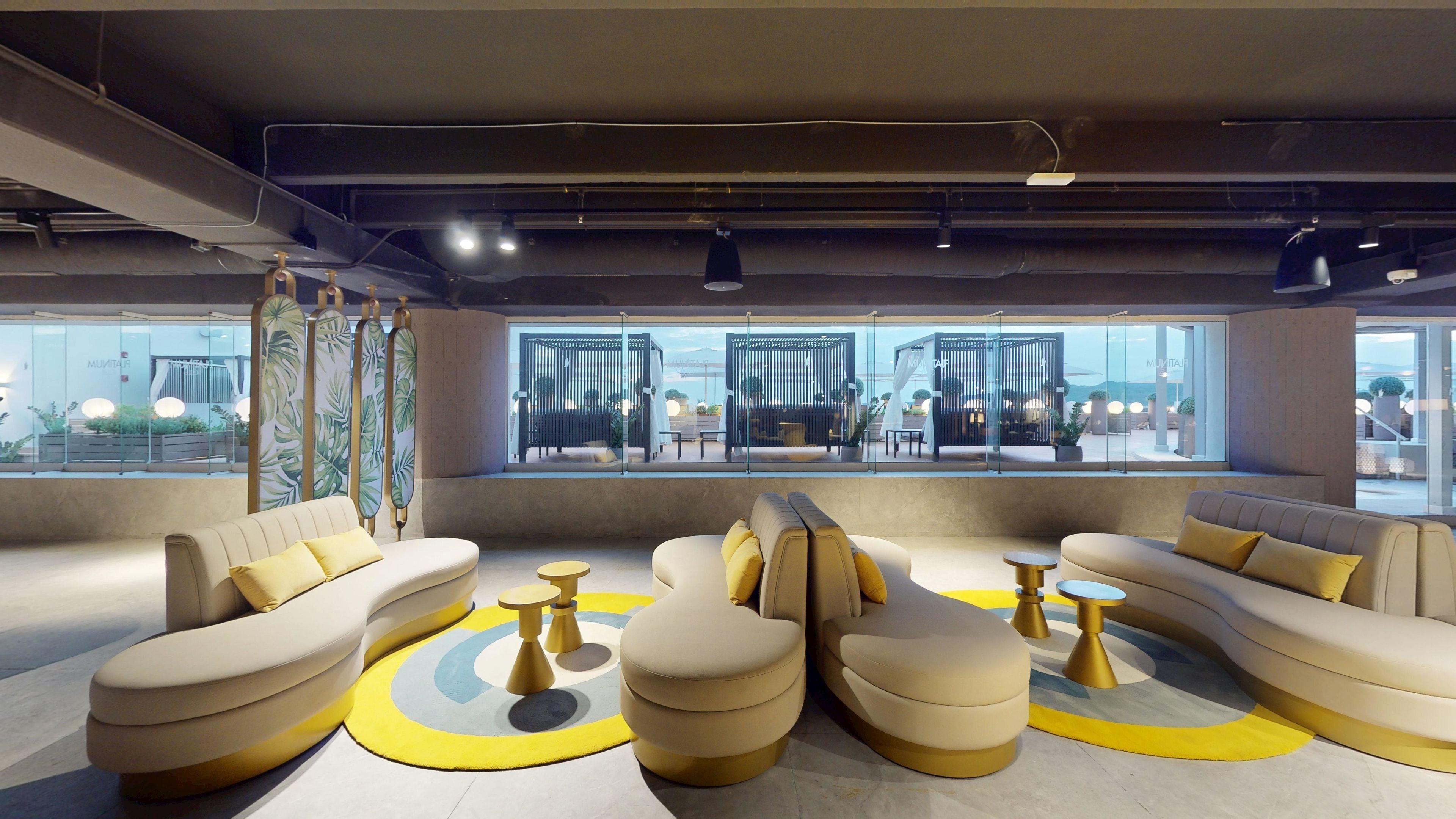 A modern lounge area with sofas and round tables, large windows offering views of the outdoors, and a ceiling with exposed ductwork.