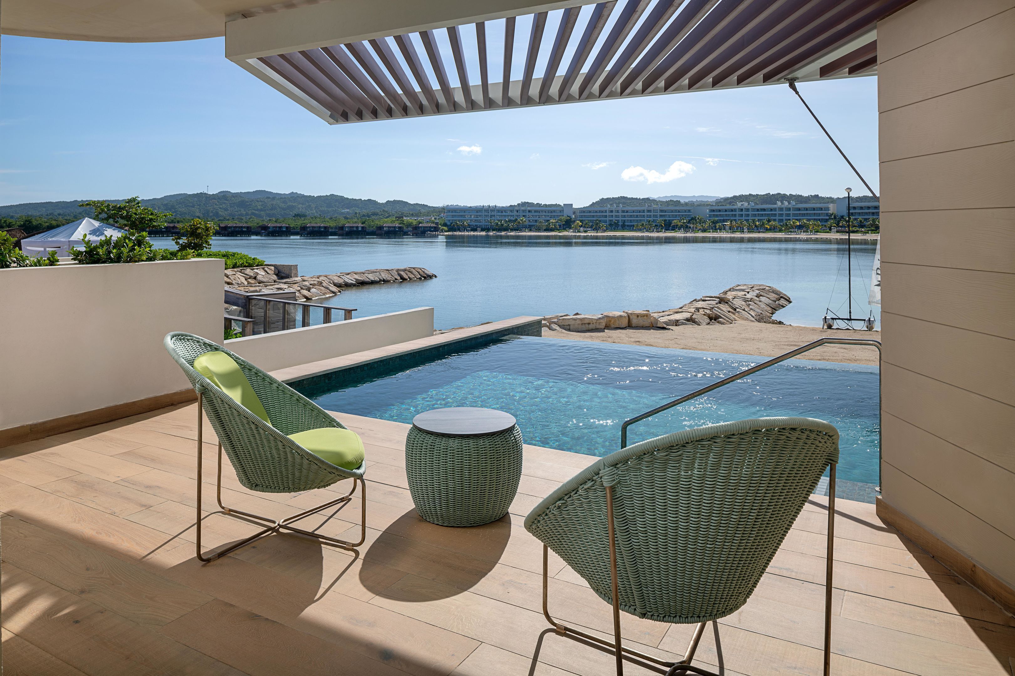 Un balcone con piscina, due poltrone in vimini e vista su un corpo d'acqua e montagne in lontananza.