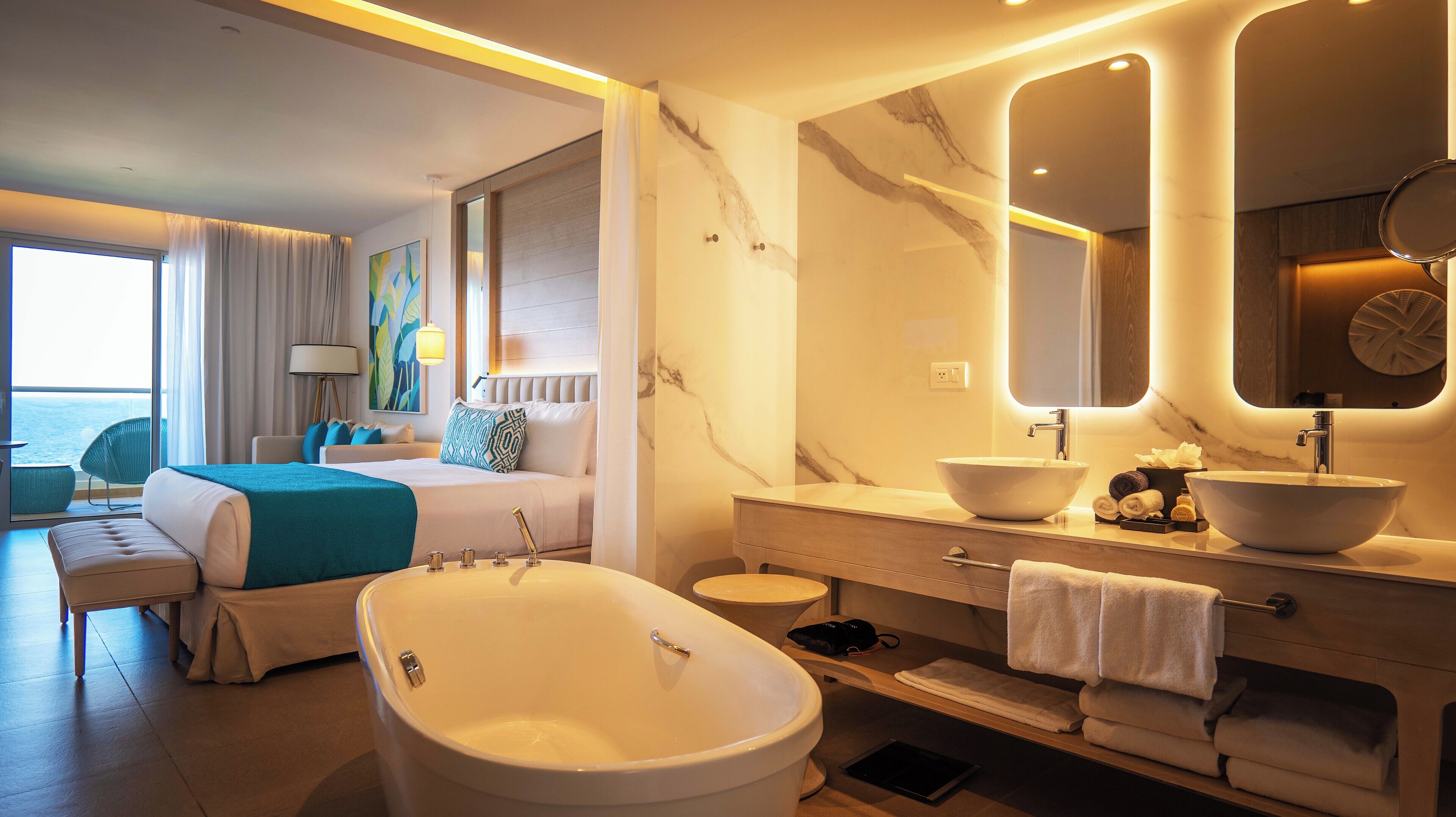 Une chambre d’hôtel moderne dotée d’un grand lit, d’un coin salon avec vue, d’une salle de bain avec une baignoire sur pieds et d’une coiffeuse avec un grand miroir.