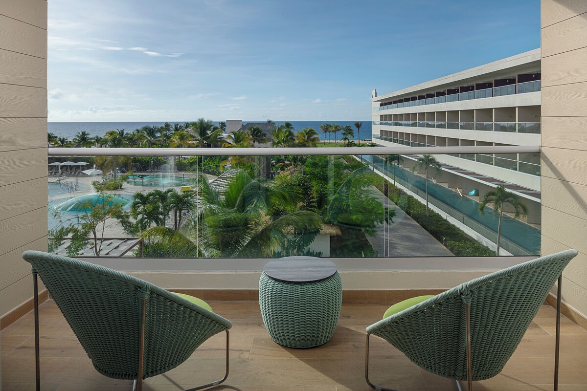 A balcony with two wicker chairs, a small round table, and a view of a swimming pool and palm trees.