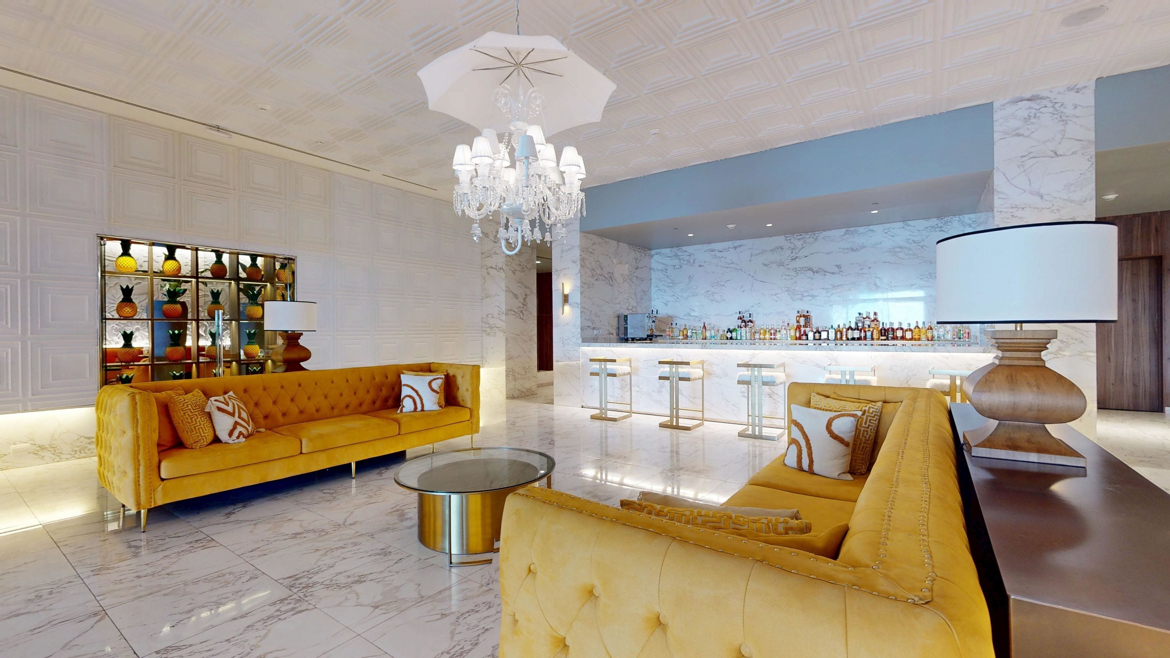 A spacious lobby with a yellow sofa, a round coffee table, a bar area with stools, and a chandelier.