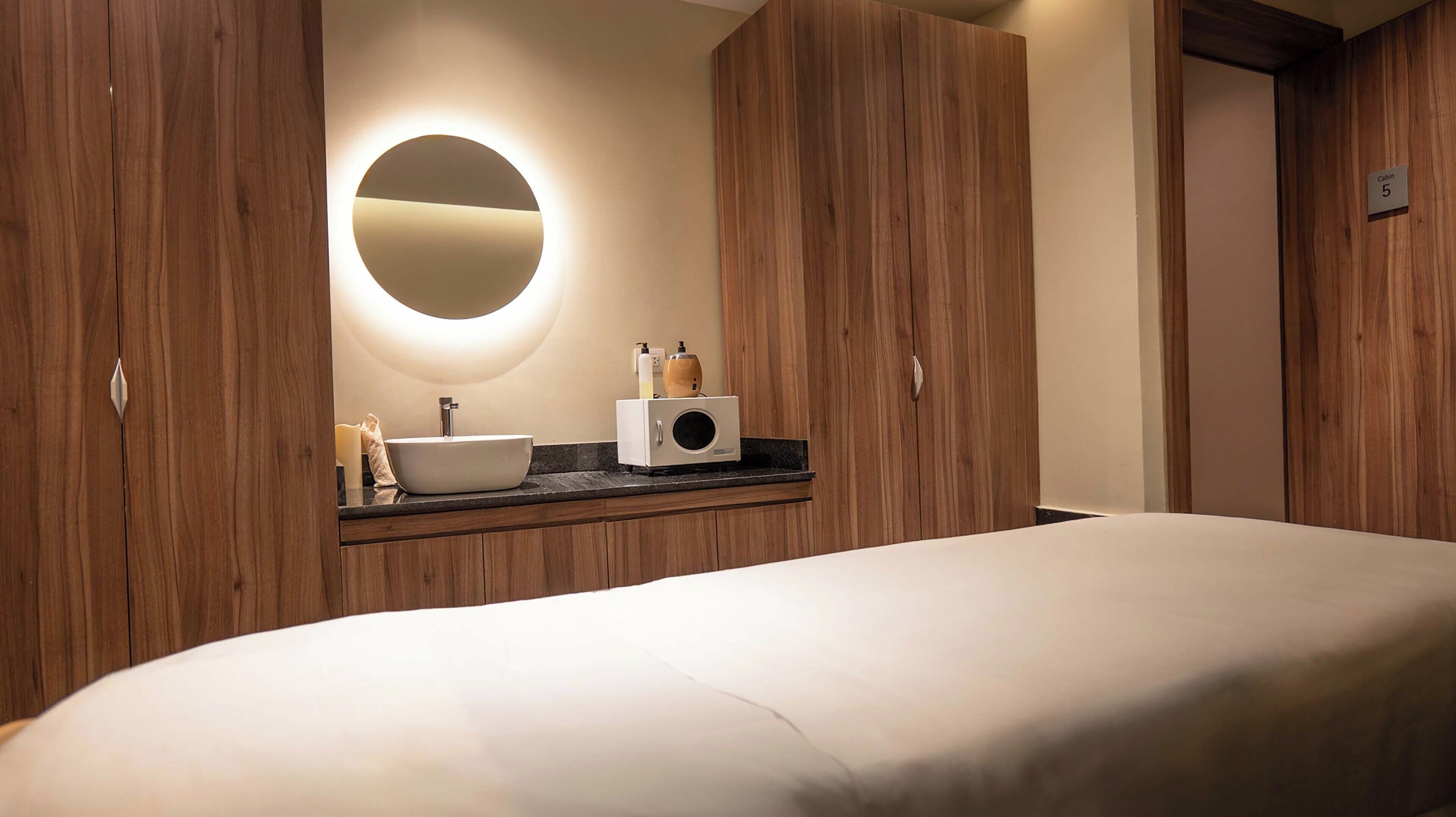 A hotel room with a bed, a round mirror, a sink, and wooden cabinets.