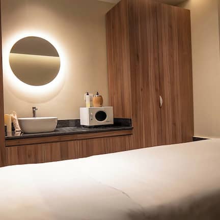 A hotel room with a bed, a round mirror, a sink, and wooden cabinets.