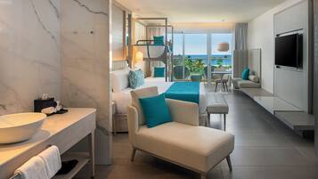 A modern hotel room with a large bathroom vanity, a sofa, a TV, and a balcony with a view.