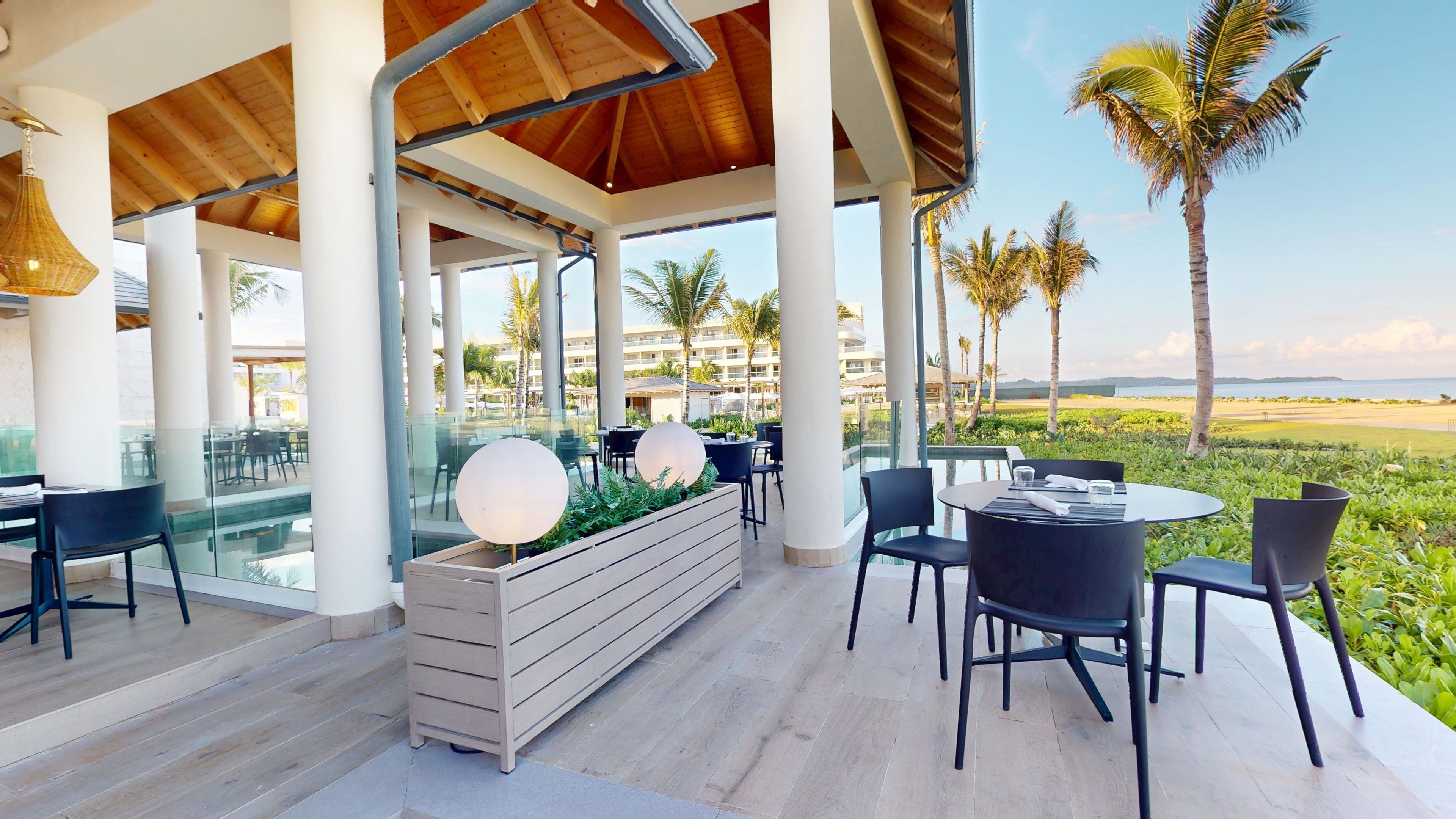 An outdoor dining area with modern furniture, a wooden planter with spherical lights, and a view of the beach and palm trees.