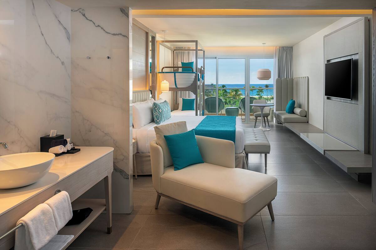 Ein modernes Hotelzimmer mit großzügigem Badezimmer-Waschtisch, einer Couch, einem Fernseher und einem Balkon mit Blick.