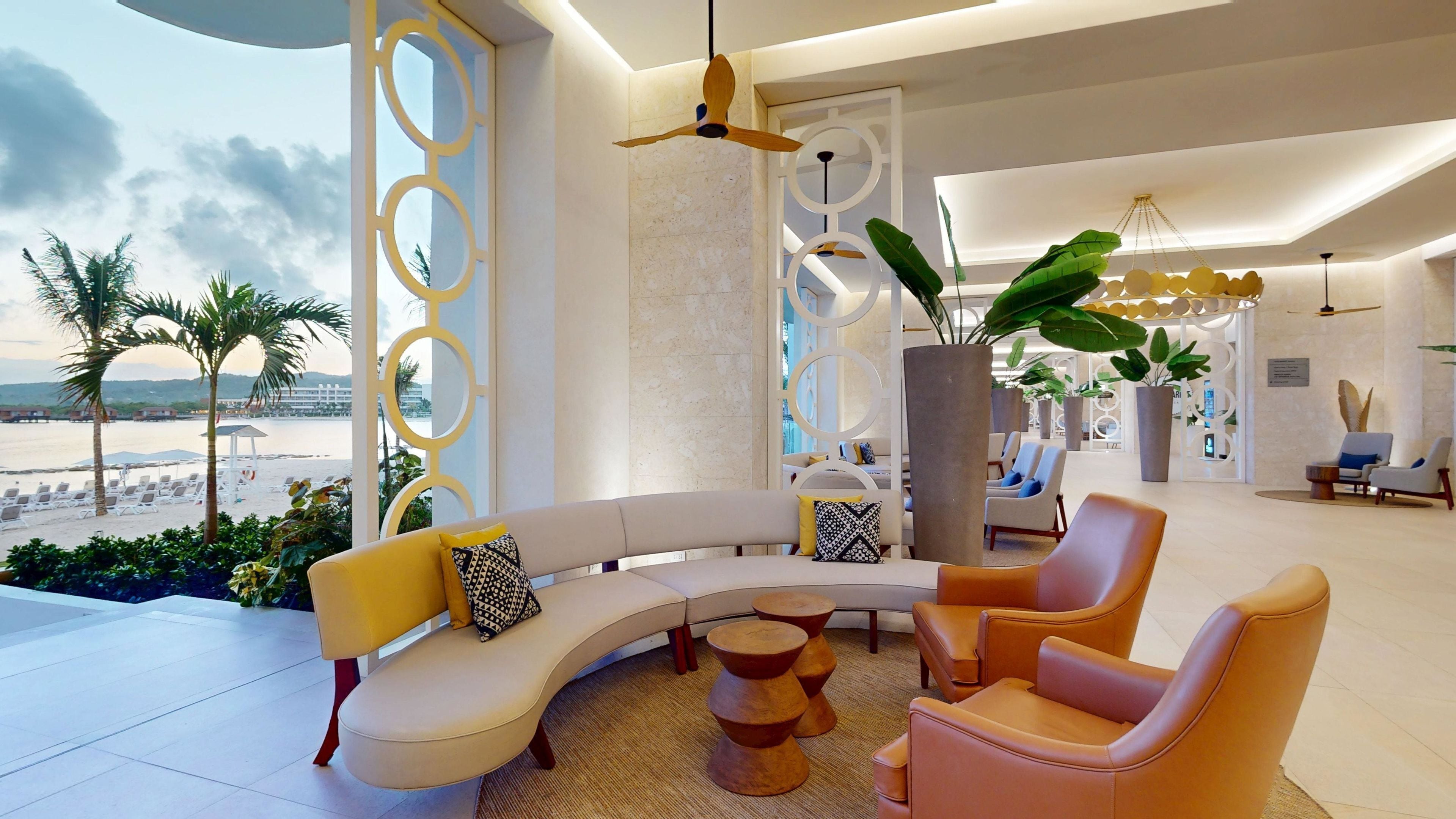 A modern lounge area with a curved sofa, two armchairs, and a coffee table. There are potted plants and a view of the beach.