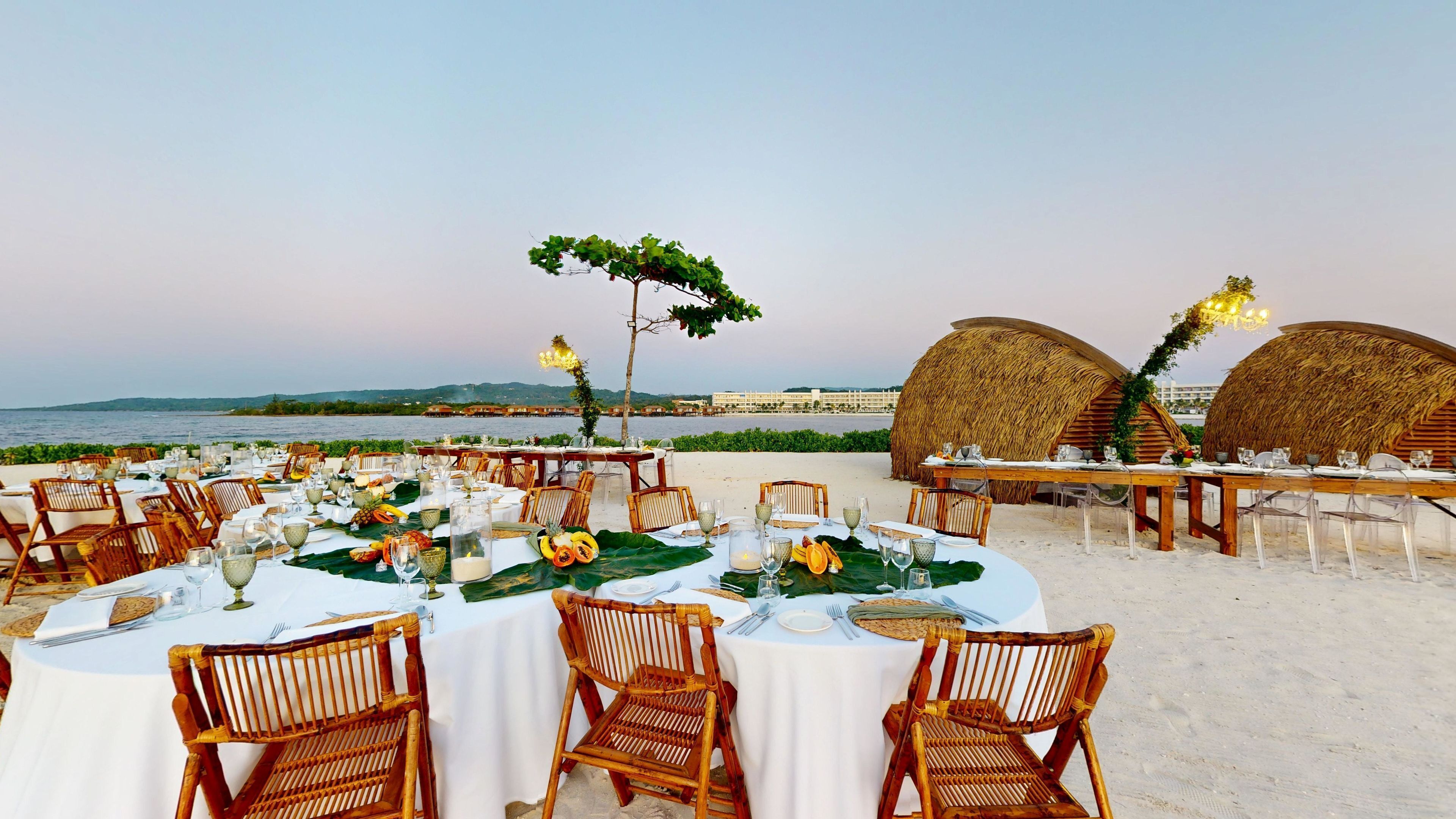 Un'area pranzo all'aperto con tavoli rotondi, sedie in vimini e vista sul mare e sugli edifici sullo sfondo.