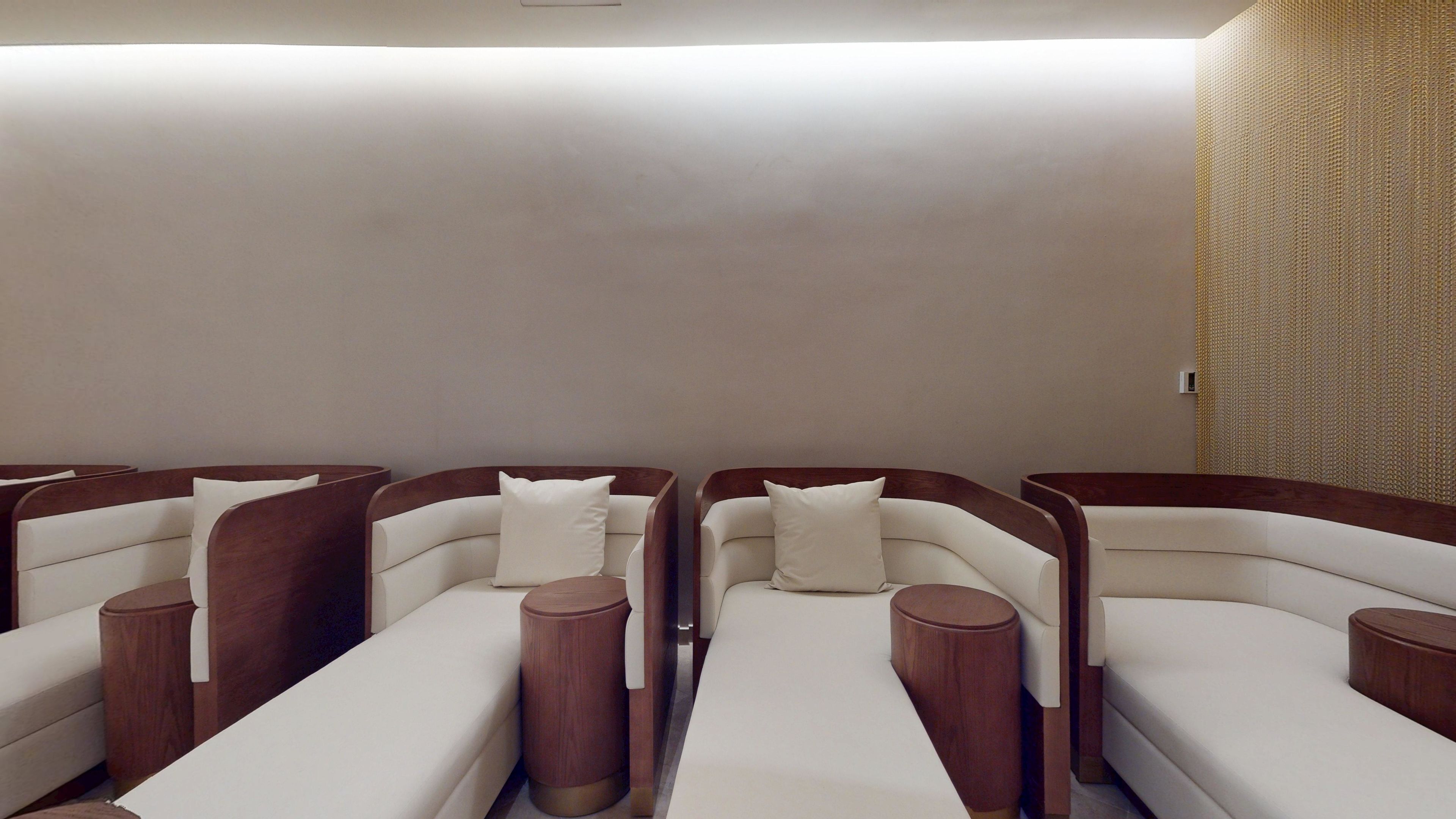 A row of four reclining chairs with wooden side panels and white cushions, arranged in a row with a plain wall and a textured wall on one side.