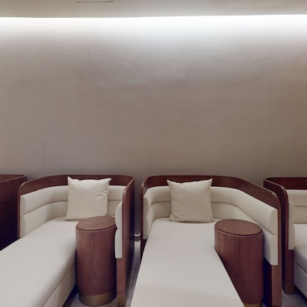 A row of four reclining chairs with wooden side panels and white cushions, arranged in a row with a plain wall and a textured wall on one side.