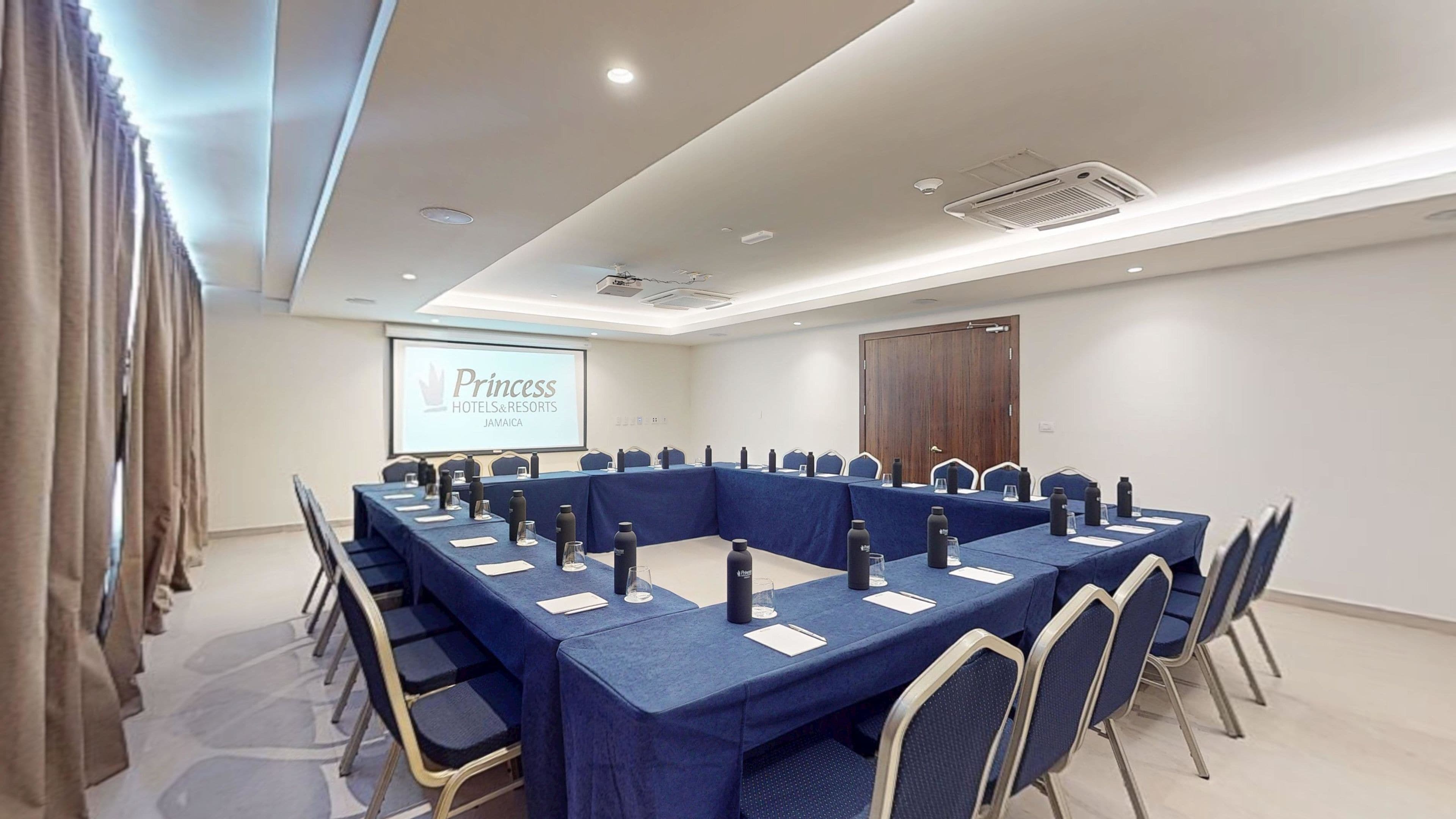 Un salón de conferencias con una larga mesa, sillas, una pantalla de proyector y una puerta de madera.