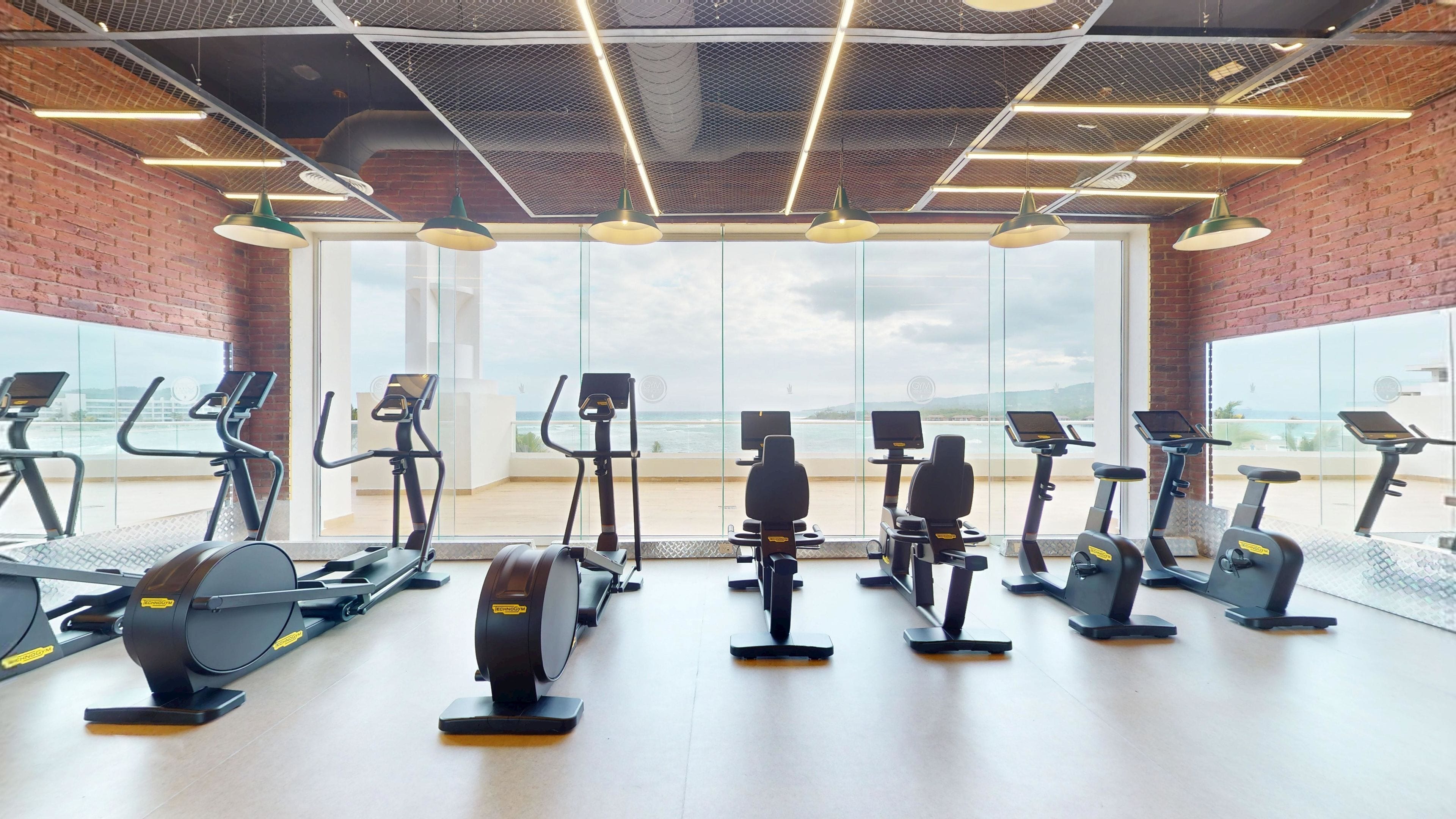 A gym with stationary bikes and treadmills, large windows with a view of the beach, and a brick wall.