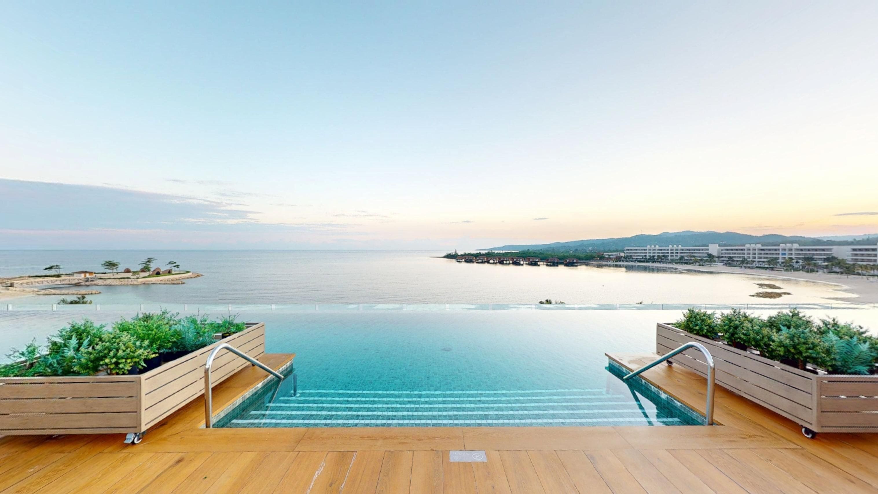 An infinity pool with wooden deck and railings, overlooking a beach and ocean.