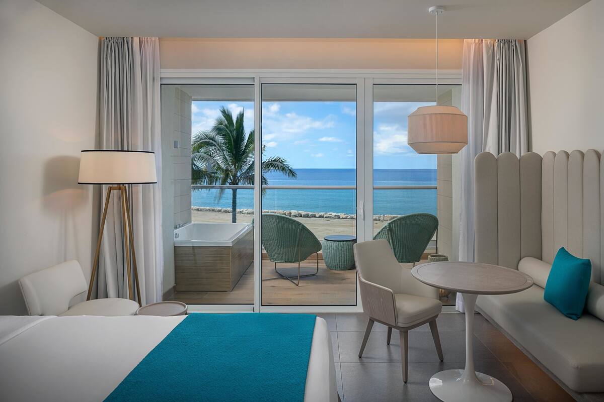 Ein Hotelzimmer mit Bett, einer Couch, einem Tisch, einem Sessel, einer Lampe und Blick auf Strand und Meer.