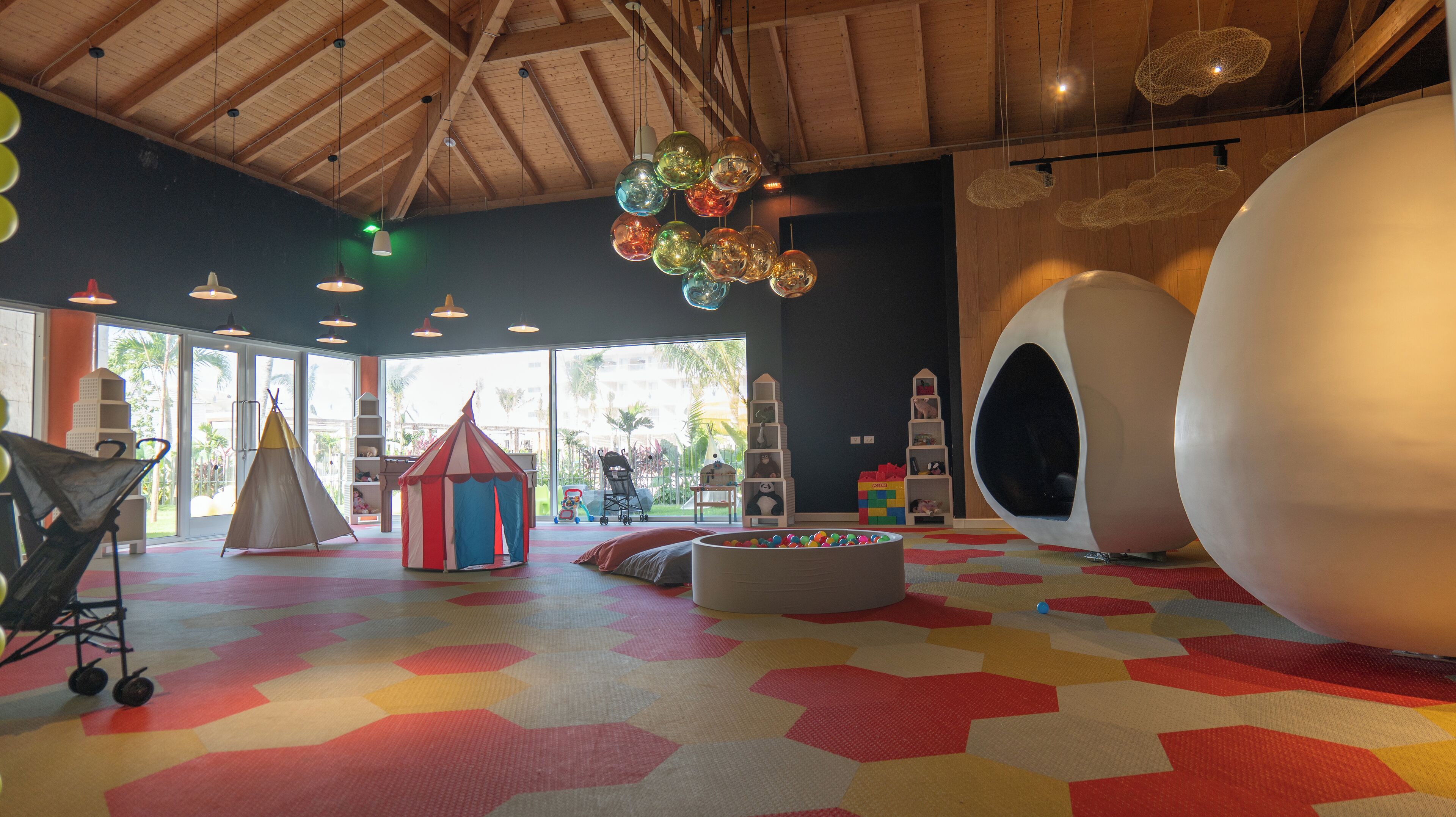 Una sala de jocs amb un paviment hexagonal de colors, una tenda de jocs, una pilota blanca gran i un llum penjant.