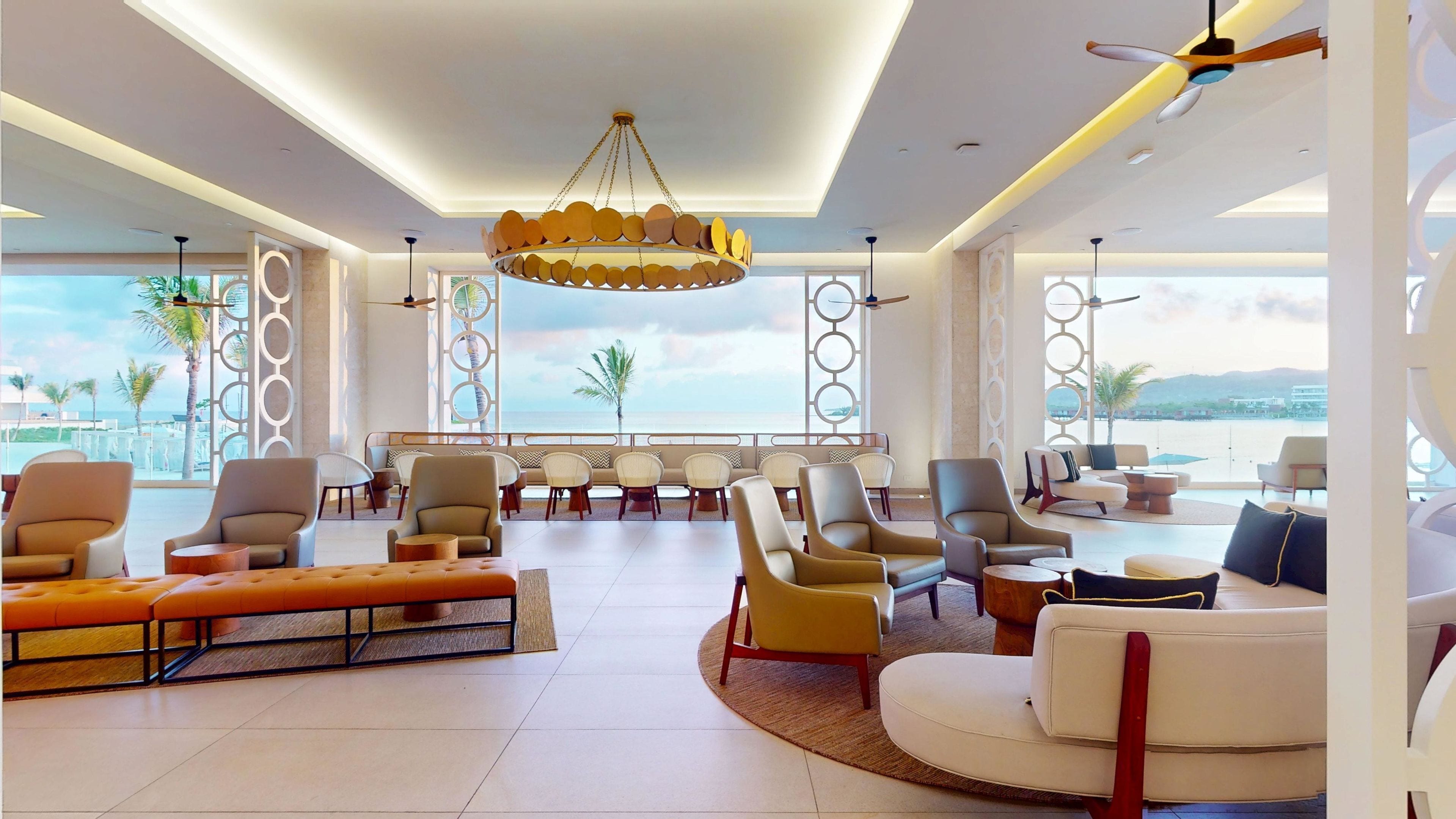 Un vaste salon aménagé avec des meubles modernes, un lustre circulaire original et de grandes fenêtres offrant une vue sur la mer.