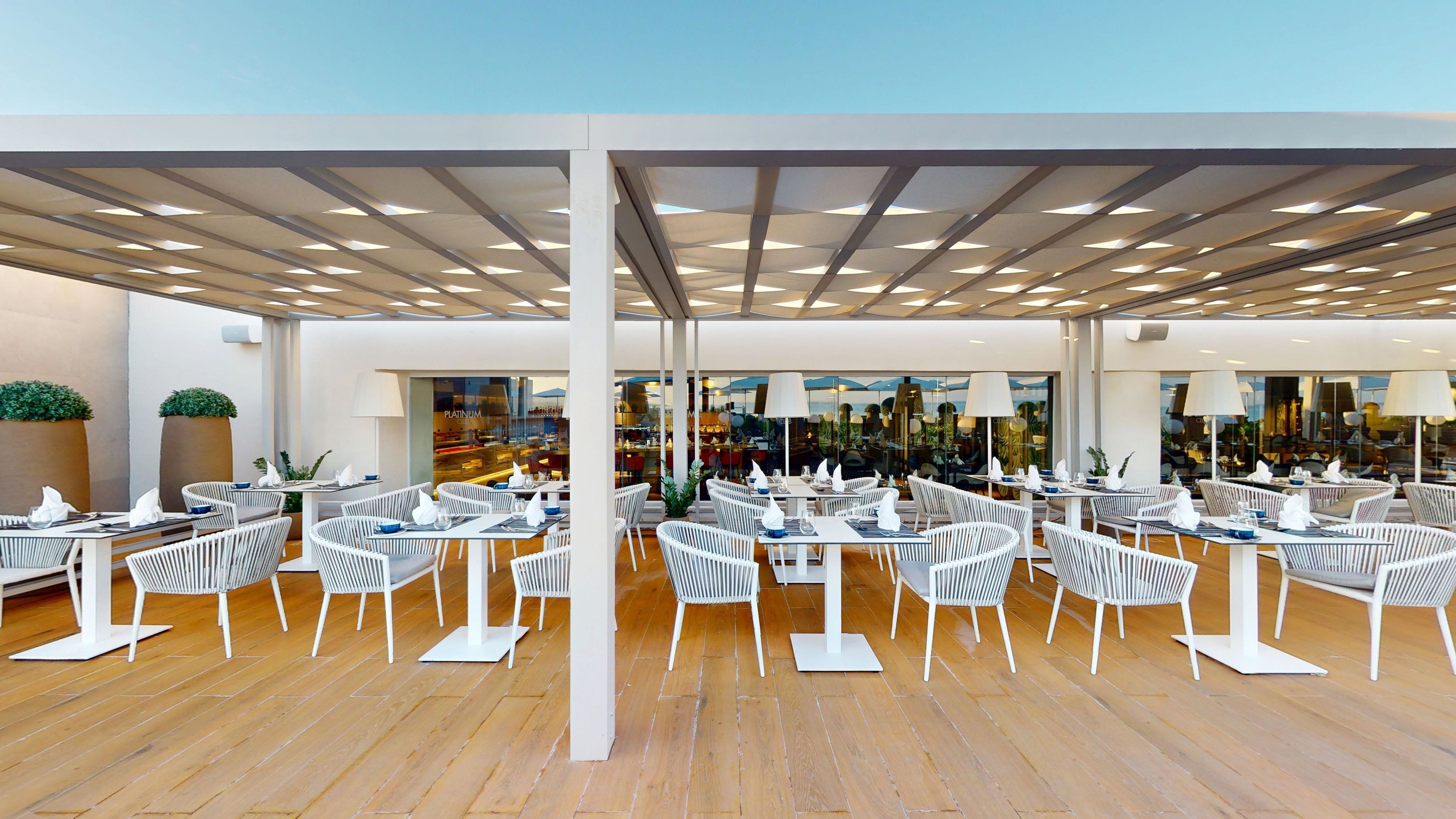 A modern restaurant with white wicker chairs and tables, wooden flooring, and a ceiling with recessed lighting.