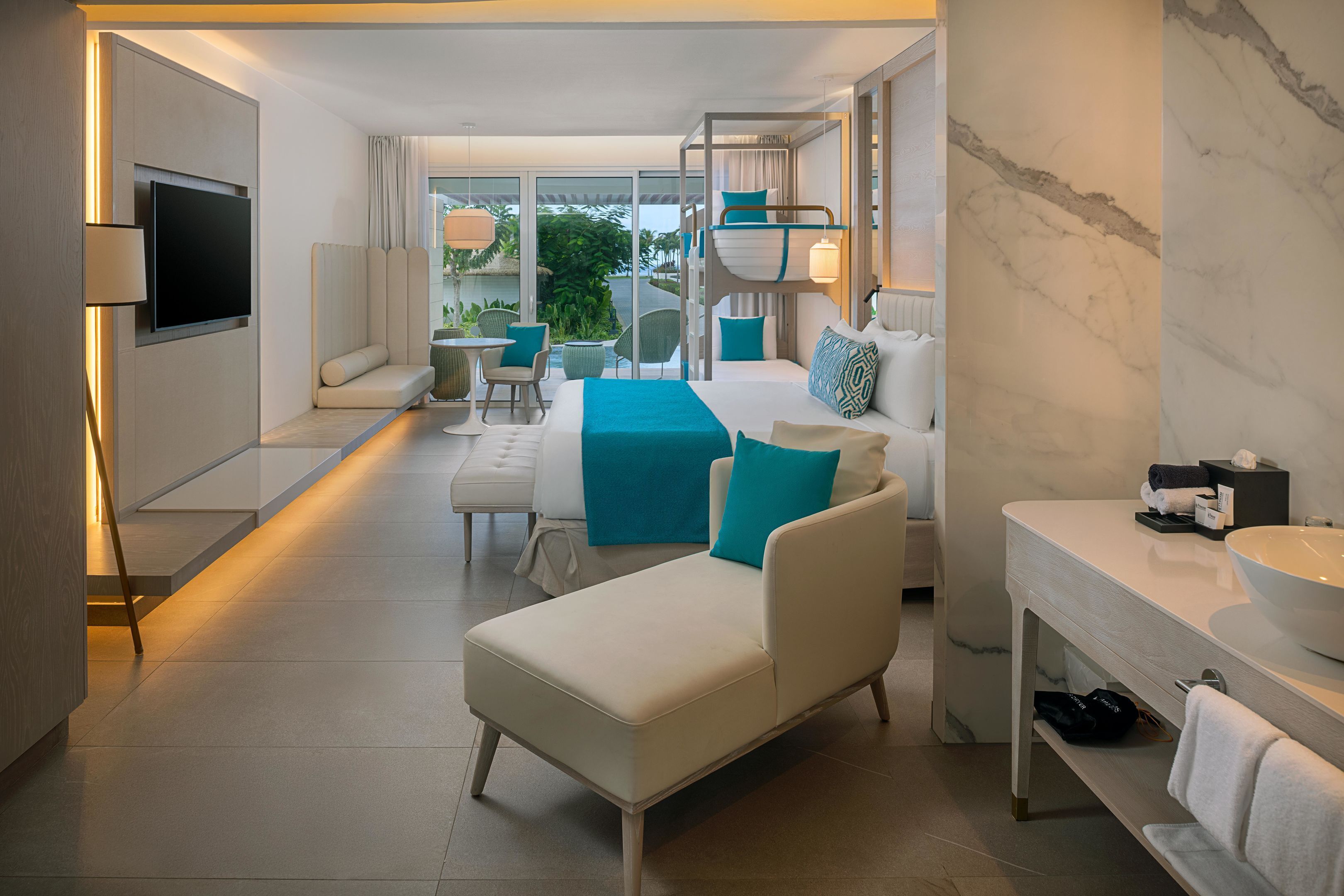 Quarto de hotel moderno com sofá, cama, televisão, varanda e banheiro com bancada de mármore.