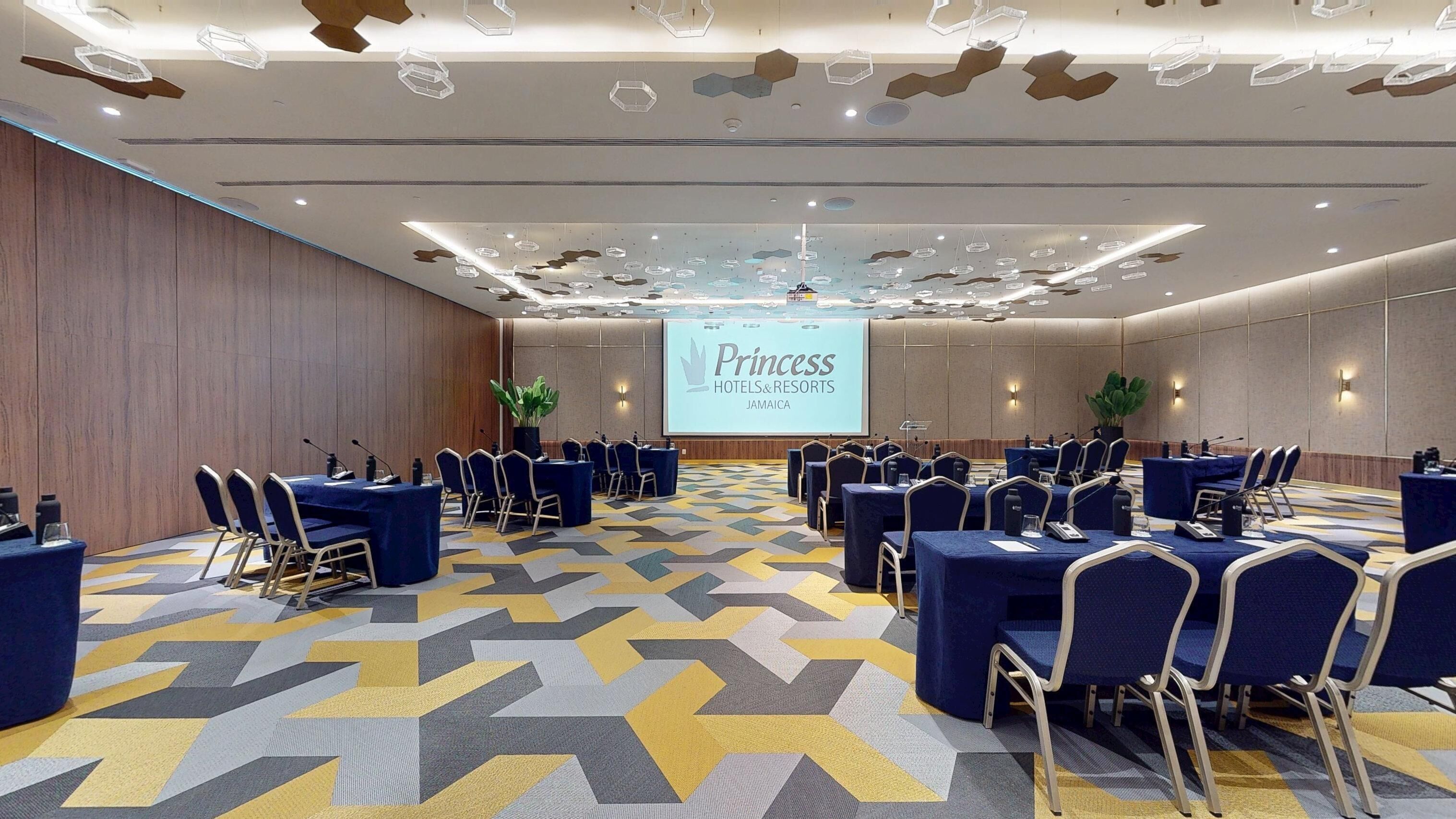 Un salón de conferencias con alfombra de estampado geométrico, filas de mesas con sillas y una pantalla grande que muestra 