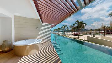 A modern outdoor pool area with a hot tub, clear blue water, and a view of the beach and palm trees.