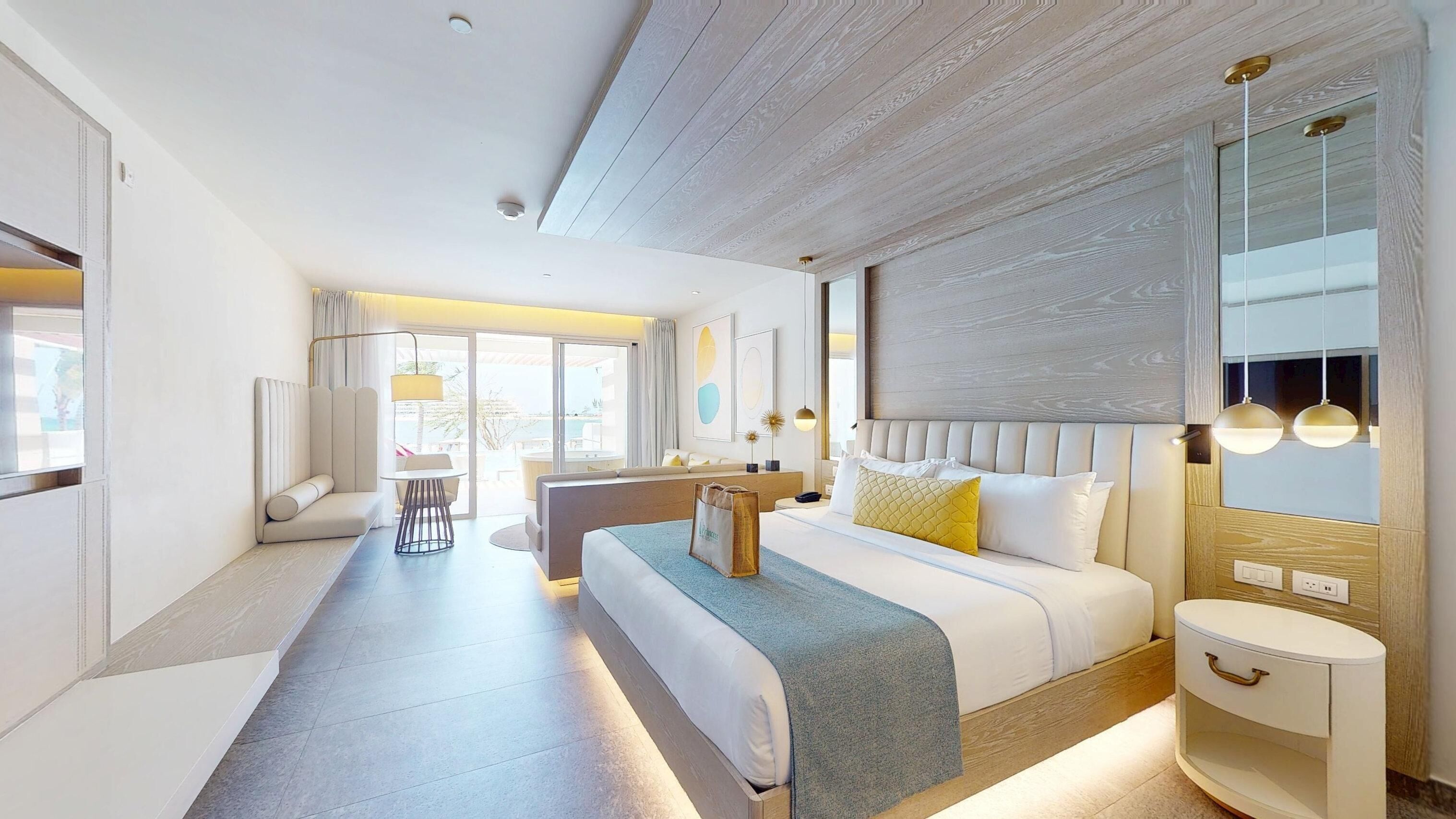 A modern hotel room with a large bed, a seating area with a sofa and armchair, a small table, and a large window offering a view.