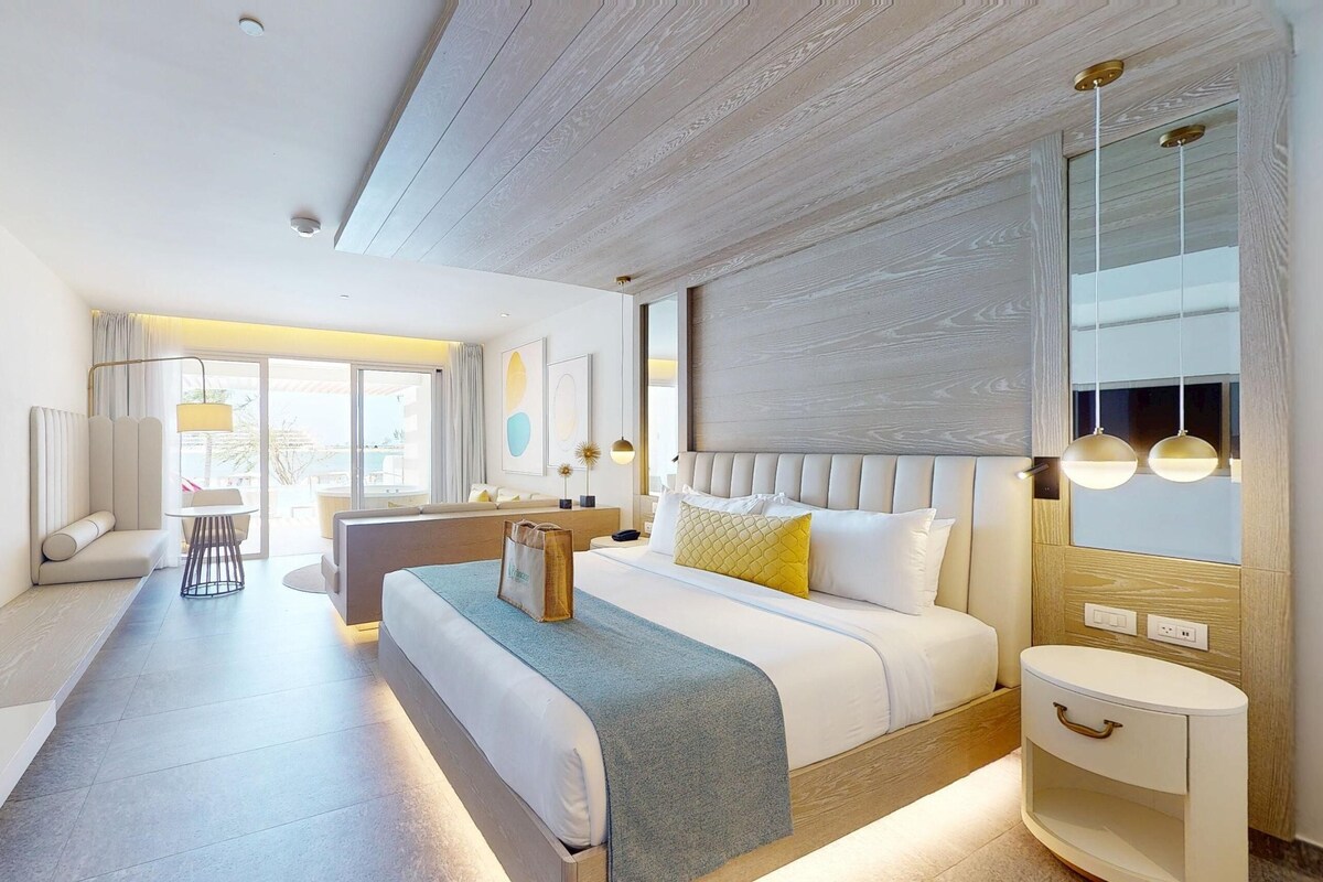 A modern hotel room with a large bed, a seating area with a sofa and armchair, a small table, and a large window offering a view.