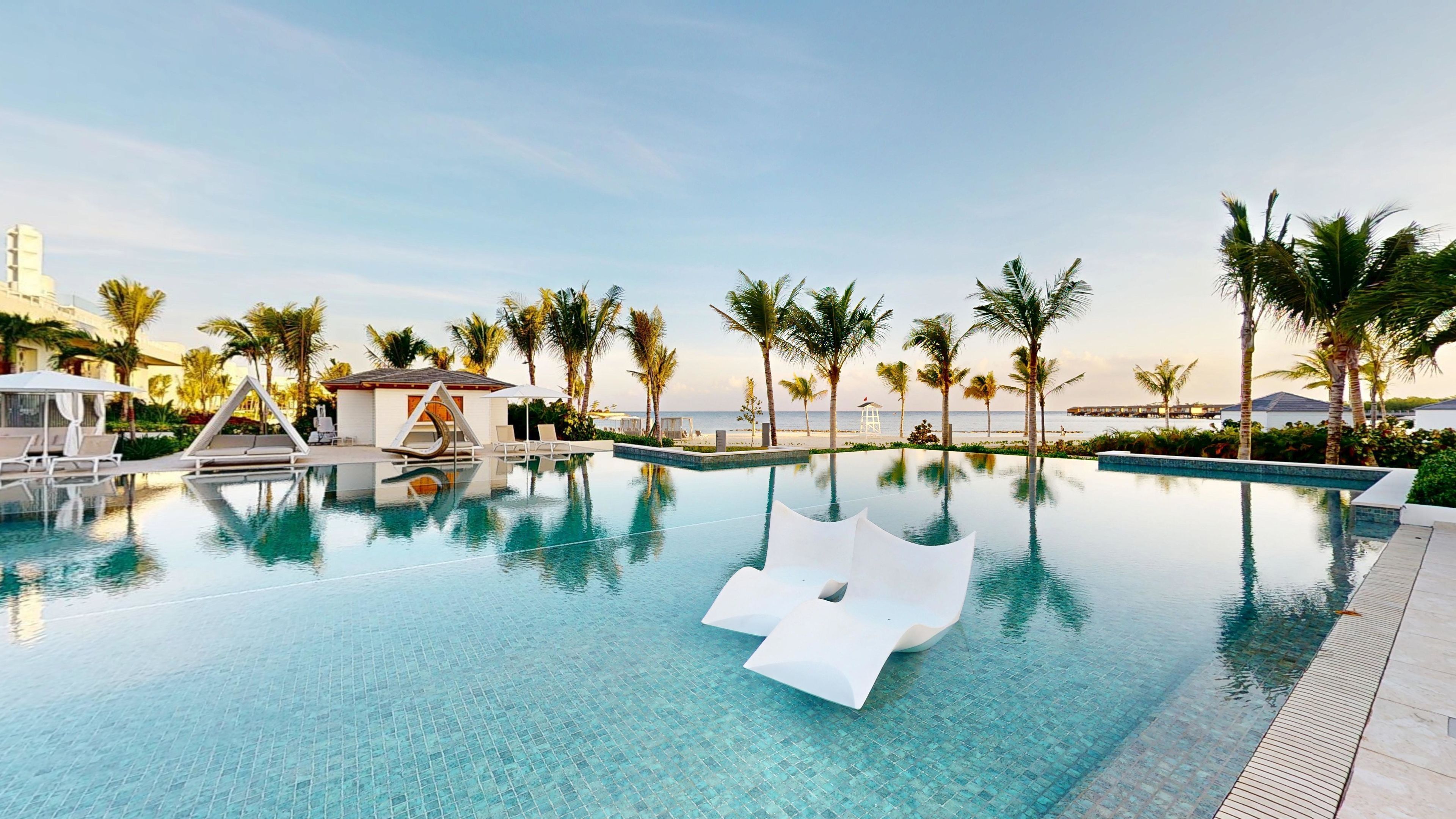 A clear swimming pool with lounge chairs and umbrellas, surrounded by palm trees and a view of the beach.