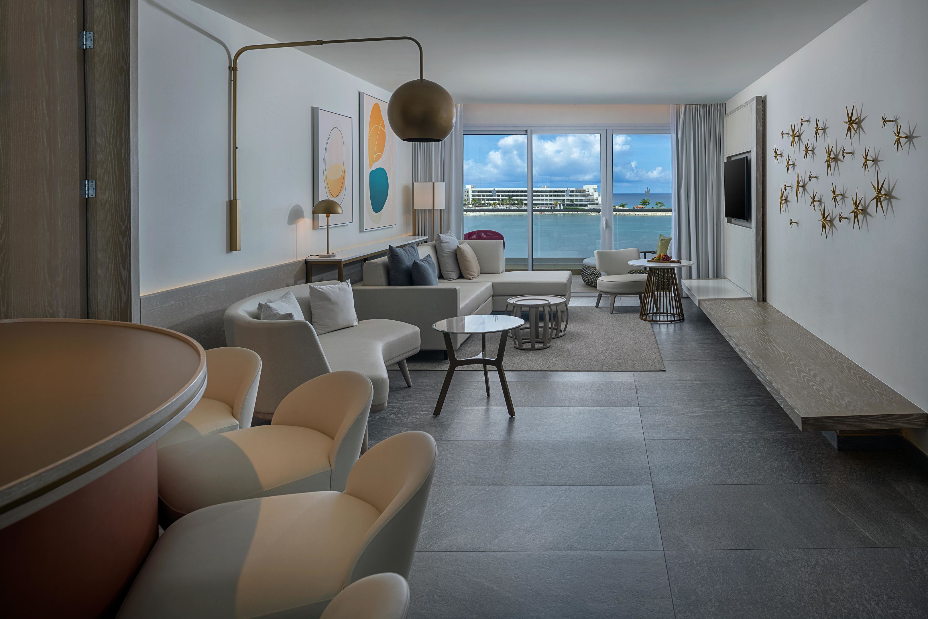 A modern hotel room with a large window, a sofa, a dining table, and a TV.
