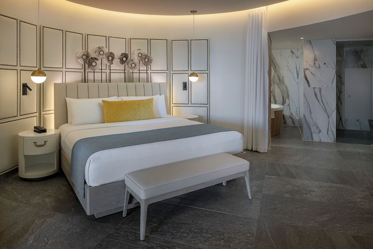 A modern bedroom with a bed, bedside tables, a bench, a wall with decorative panels, and a bathroom with a marble wall and shower.
