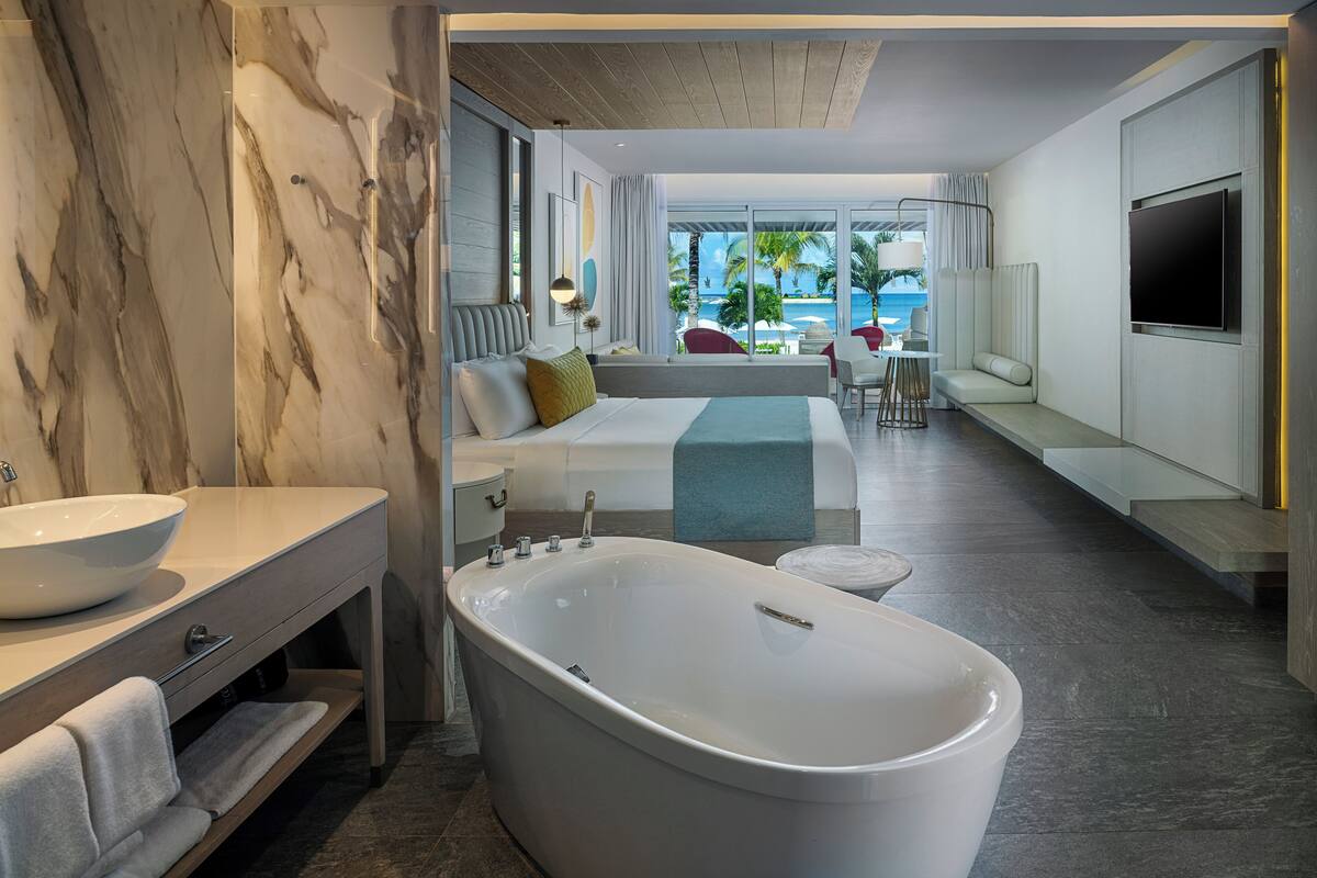 A modern hotel room with a large bathtub, a sofa bed, a dining area with a view, and a TV mounted on the wall.