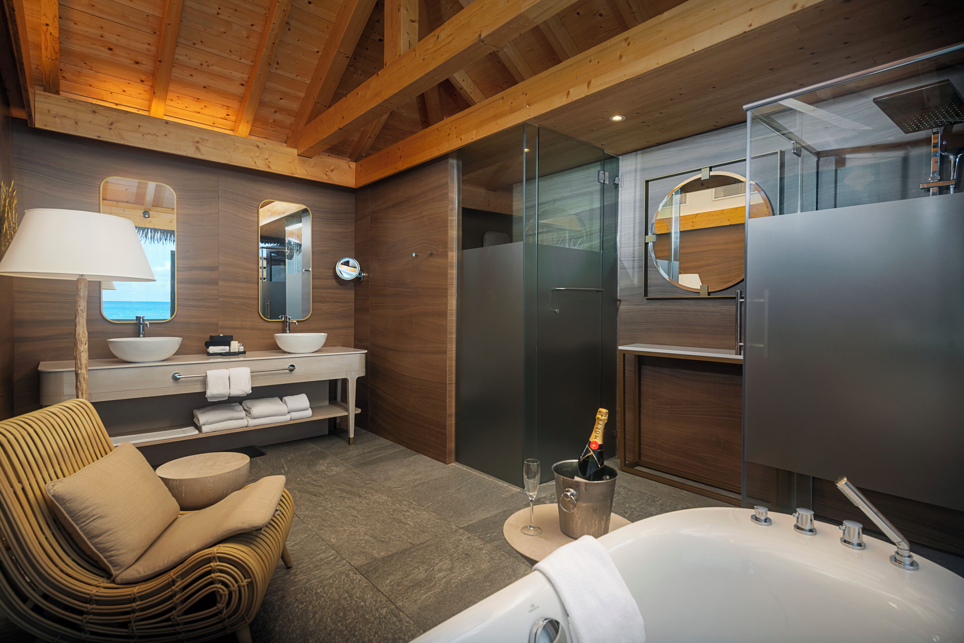 A modern bathroom with a bathtub, a glass-enclosed shower, a vanity with a sink and mirror, a wicker chair, and a bottle of champagne.
