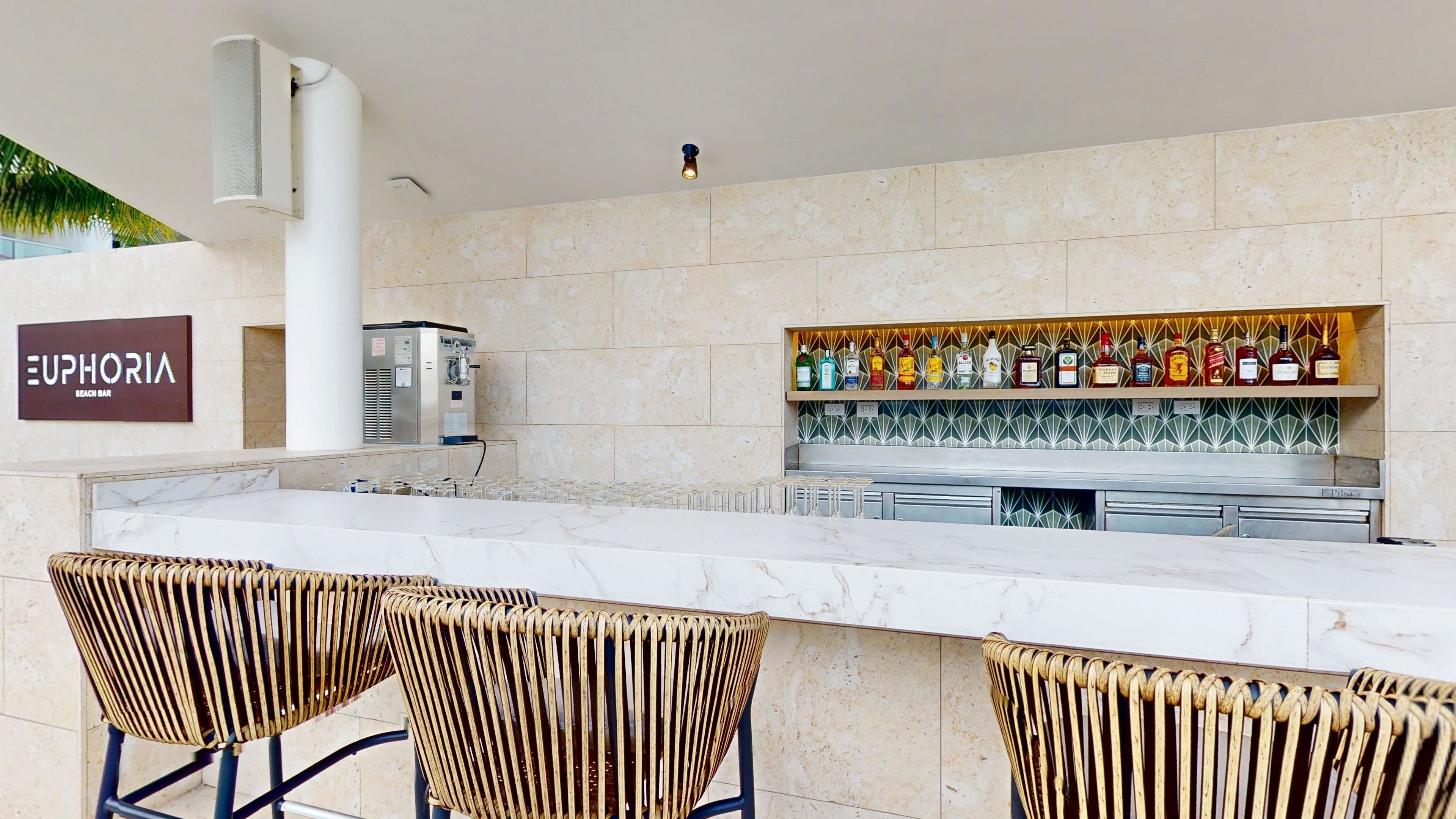 Zona de bar con encimera de mármol, sillas de mimbre, un minibar y un estante con botellas de licores.
