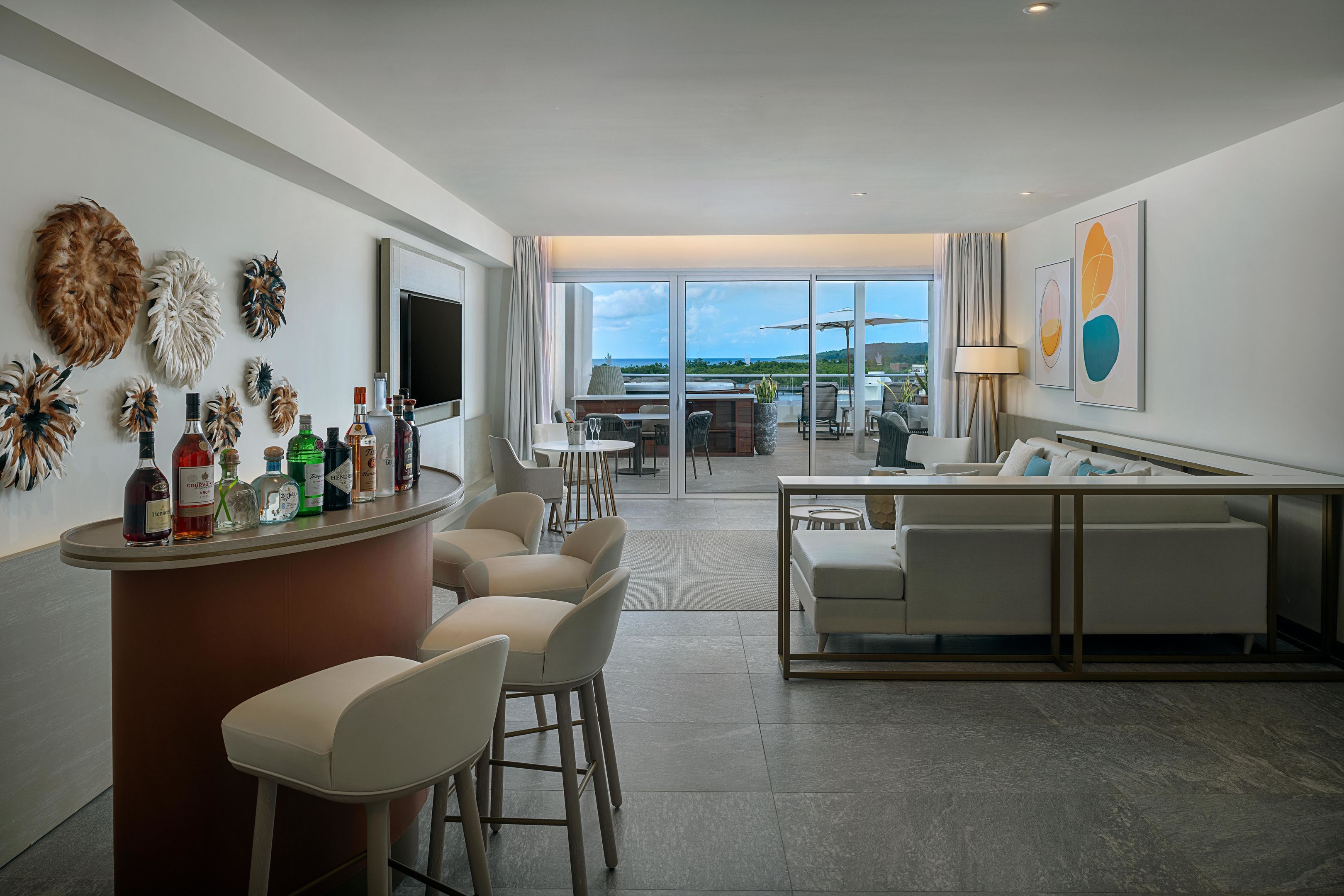 A modern hotel room with a bar area, seating, a television, and a view of the ocean.