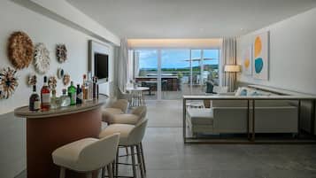 A modern hotel room with a bar area, seating, a television, and a view of the ocean.