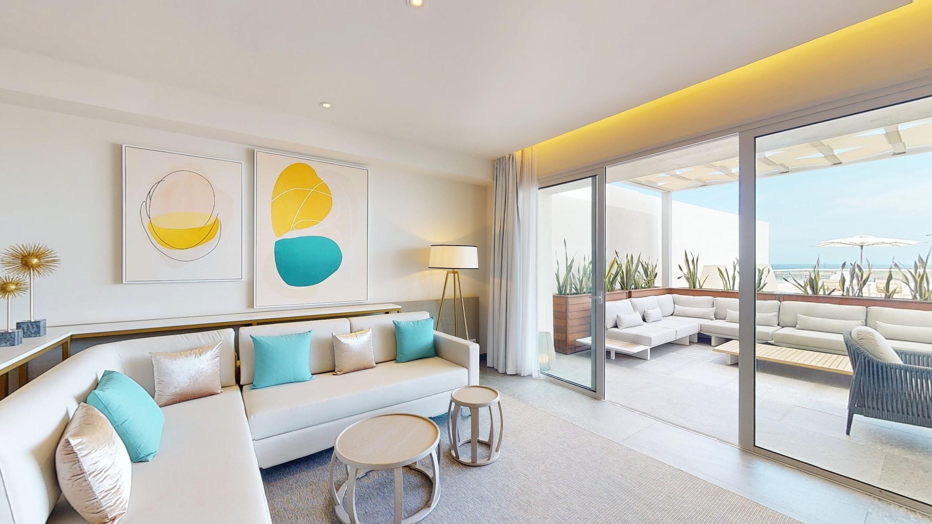 A modern living room with a sofa, two round tables, and a large sliding glass door leading to an outdoor seating area with a view of the ocean.