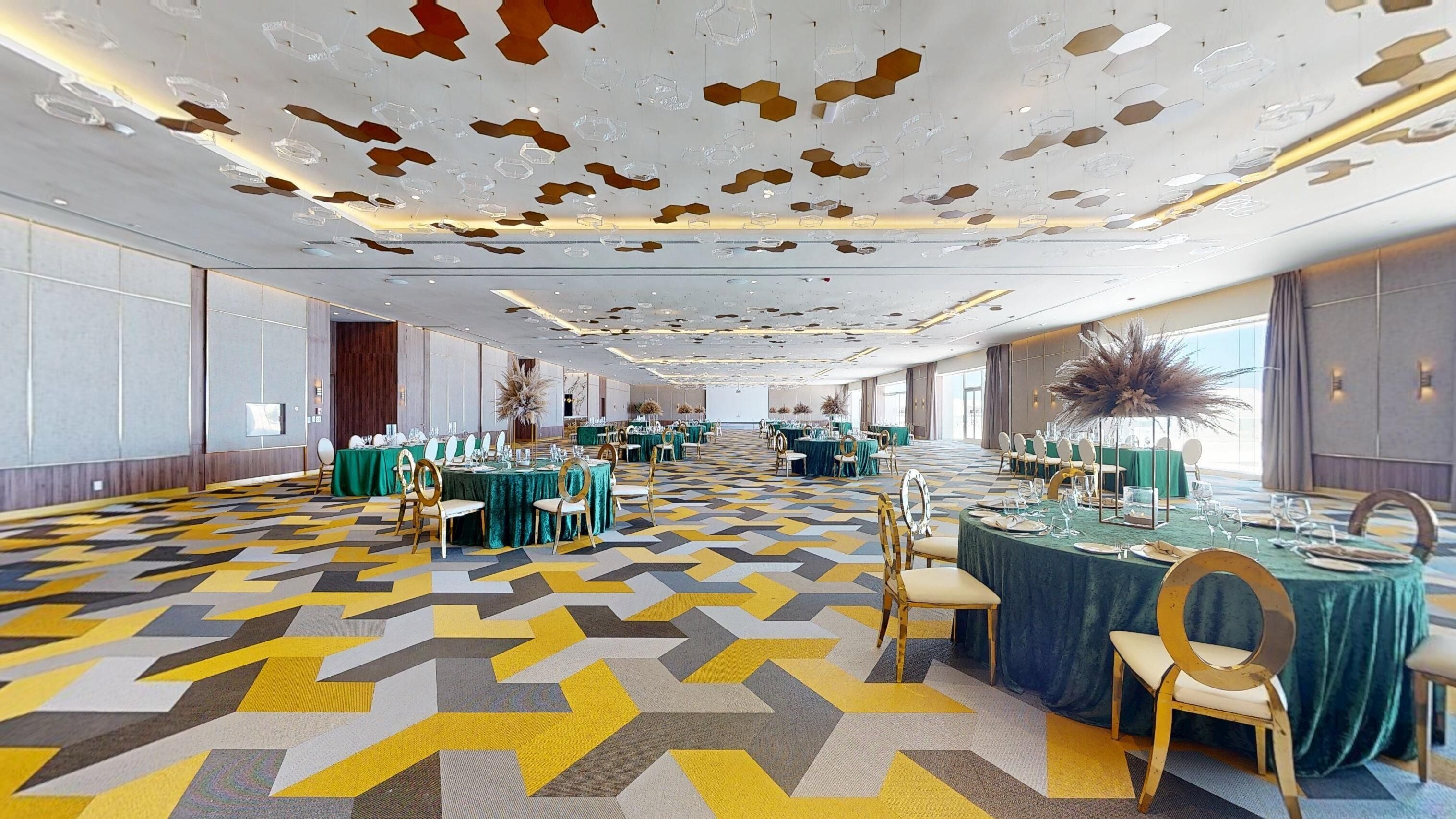 Un amplio salón de banquetes con piso de diseño geométrico, mesas redondas con manteles verdes y sillas doradas.