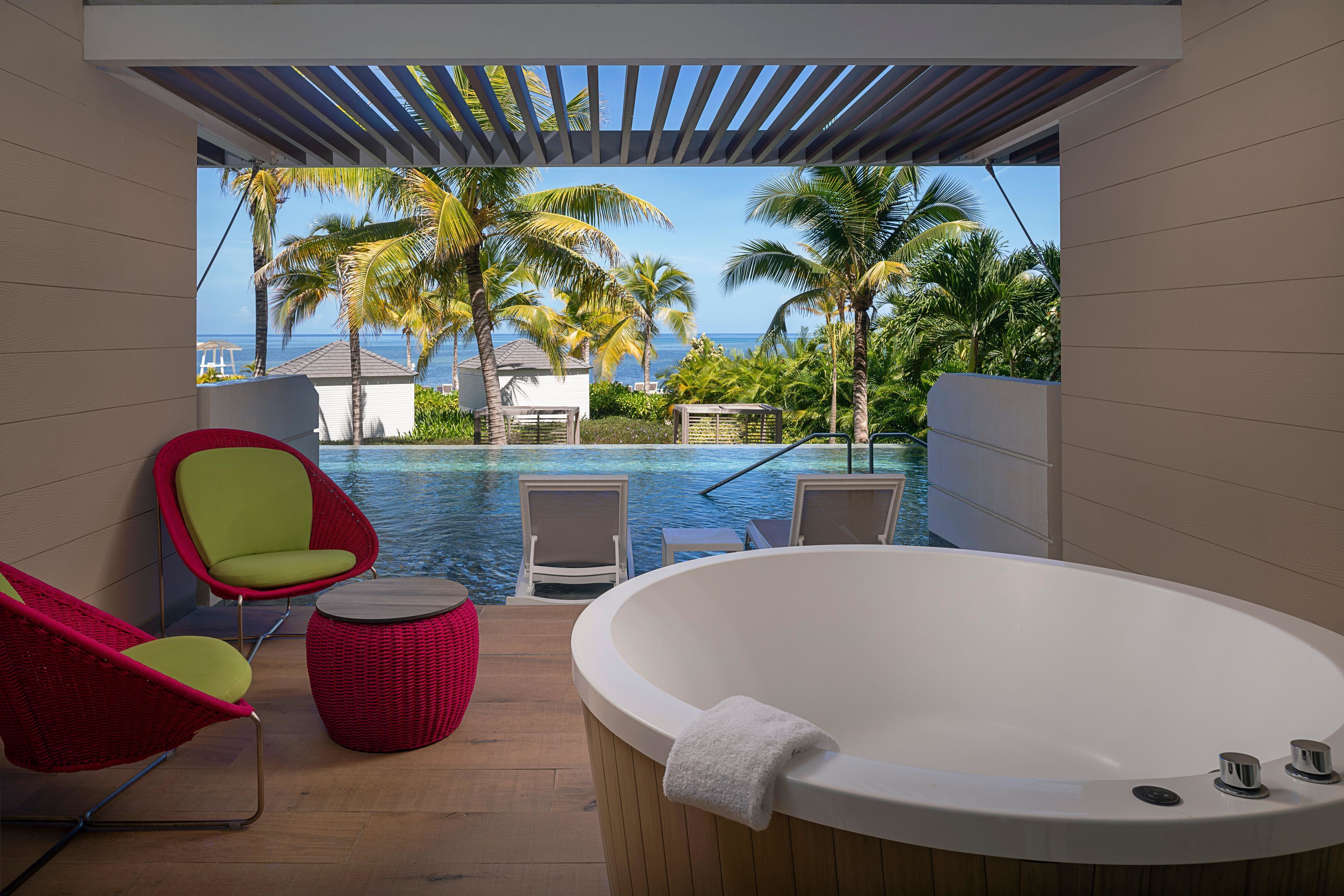 Une salle de bain moderne avec une grande baignoire, un fauteuil rouge et une vue sur une piscine et des palmiers.