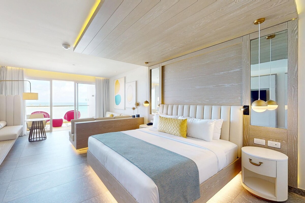 A modern hotel room with a large bed, a seating area with a sofa and chairs, a small table, and a view of the sea through the sliding glass doors.
