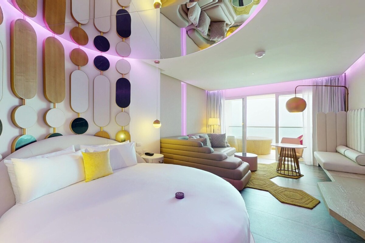 A modern hotel room with a large bed, a sofa, a small table, and a balcony with a view.
