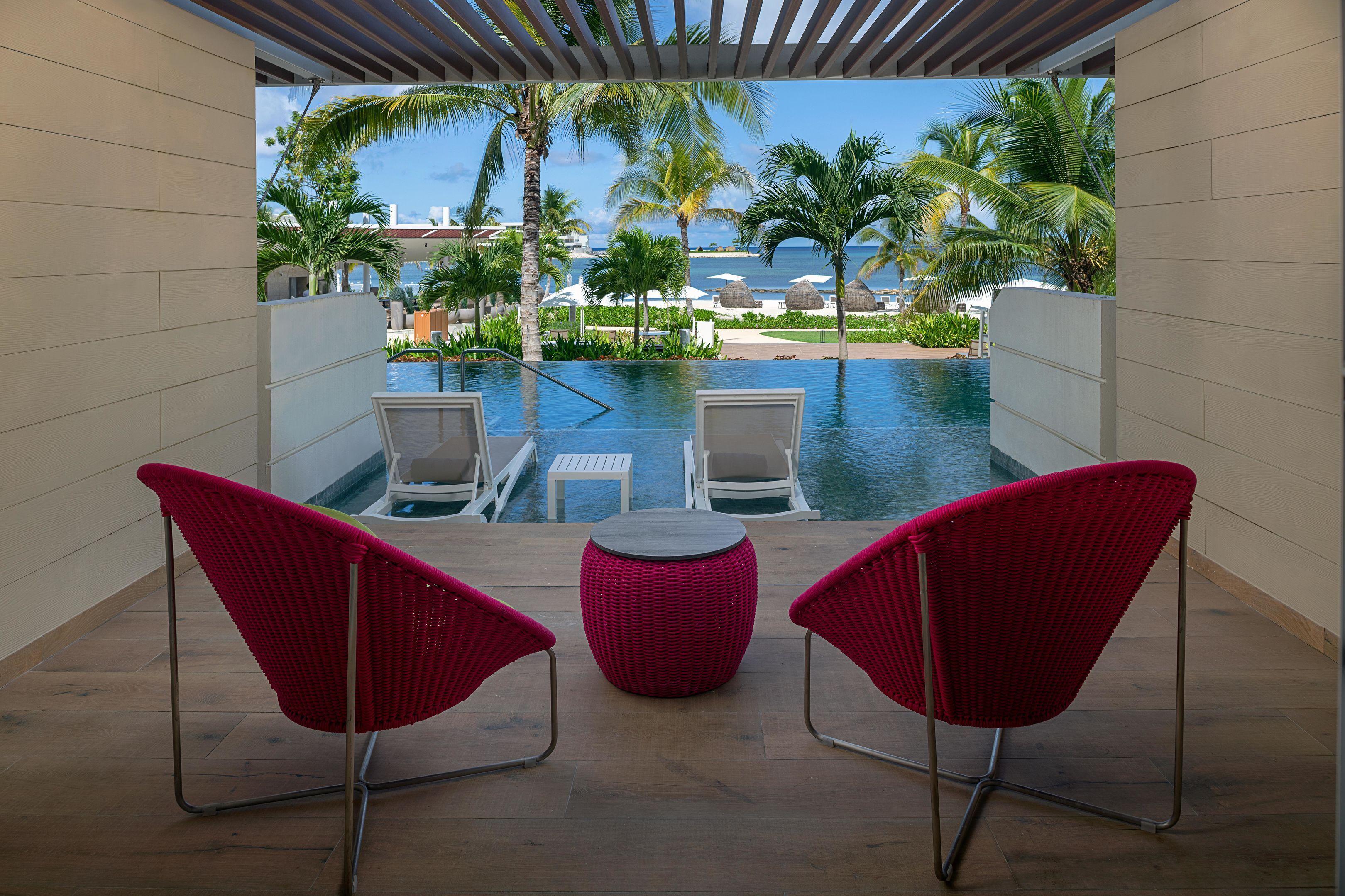 A poolside area with two red chairs, a round table, and a view of the ocean and palm trees.