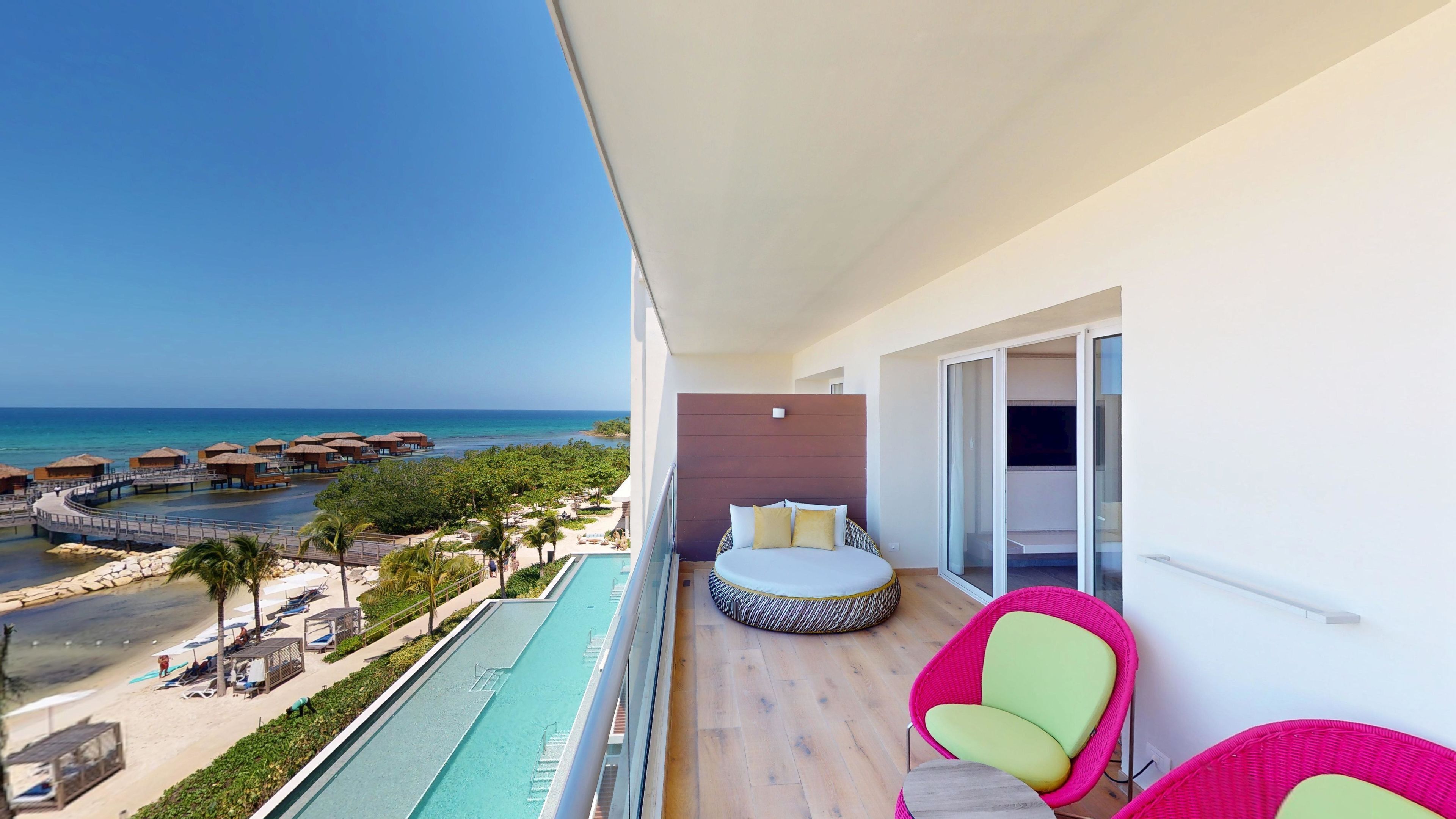 Un balcon avec un lit, deux chaises et une vue sur la plage et l’océan.