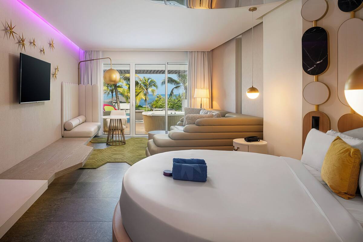 A modern hotel room with a large bed, a flat-screen TV, a sofa, a dining area with a view, and a balcony with palm trees.