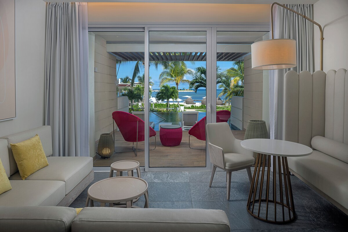 Quarto de hotel moderno com sofá, poltronas, uma mesa pequena, uma luminária e vista para uma piscina e palmeiras.