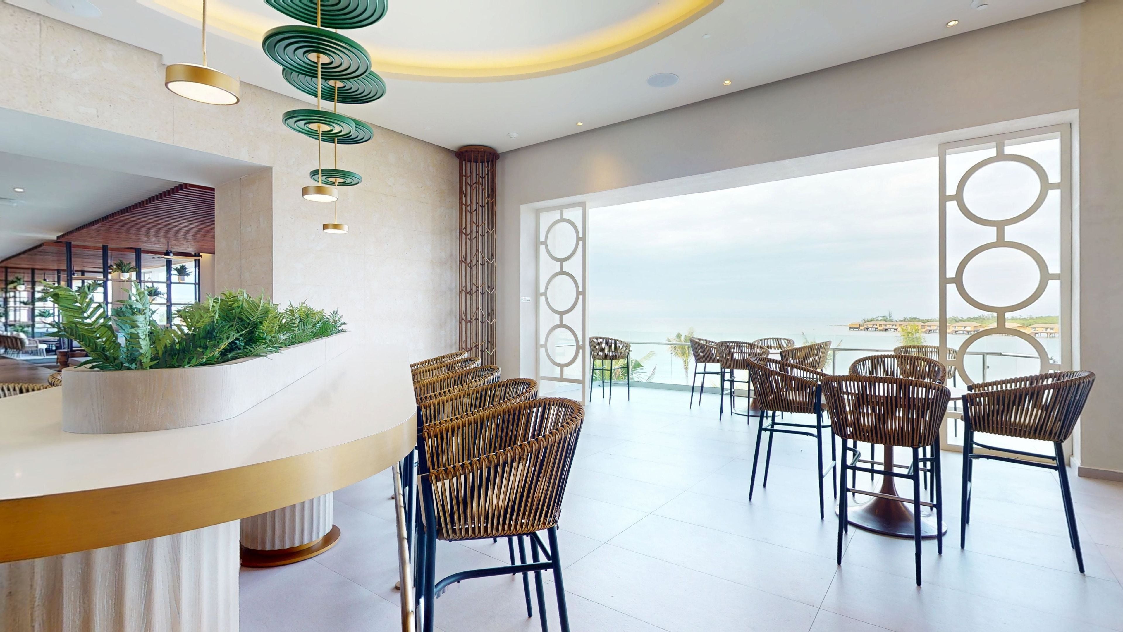 Un hall d’hôtel moderne avec un comptoir d’accueil incurvé, des fauteuils en osier et une vue sur la mer.