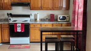 A kitchen with wooden cabinets, a white stove, a microwave, and a red kettle. There are two wooden stools at a table. - Vacation Condo #2 2BR with full kitchen, walk to river and shops, pets welcome (Hardy)