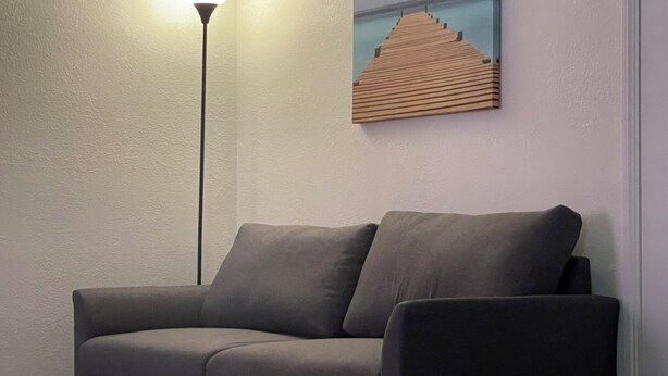 A grey sofa against a textured wall with a painting of a wooden pier extending into a body of water. A tall floor lamp with a white shade is positioned to the left of the sofa. - Vacation Condo #2 2BR with full kitchen, walk to river and shops, pets welcome (Hardy)